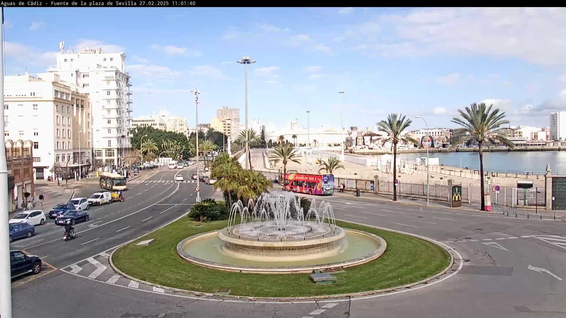 A traffic circle with a fountain in the center is surrounded by roadways, buildings, palm trees, and a body of water under a sunny sky.