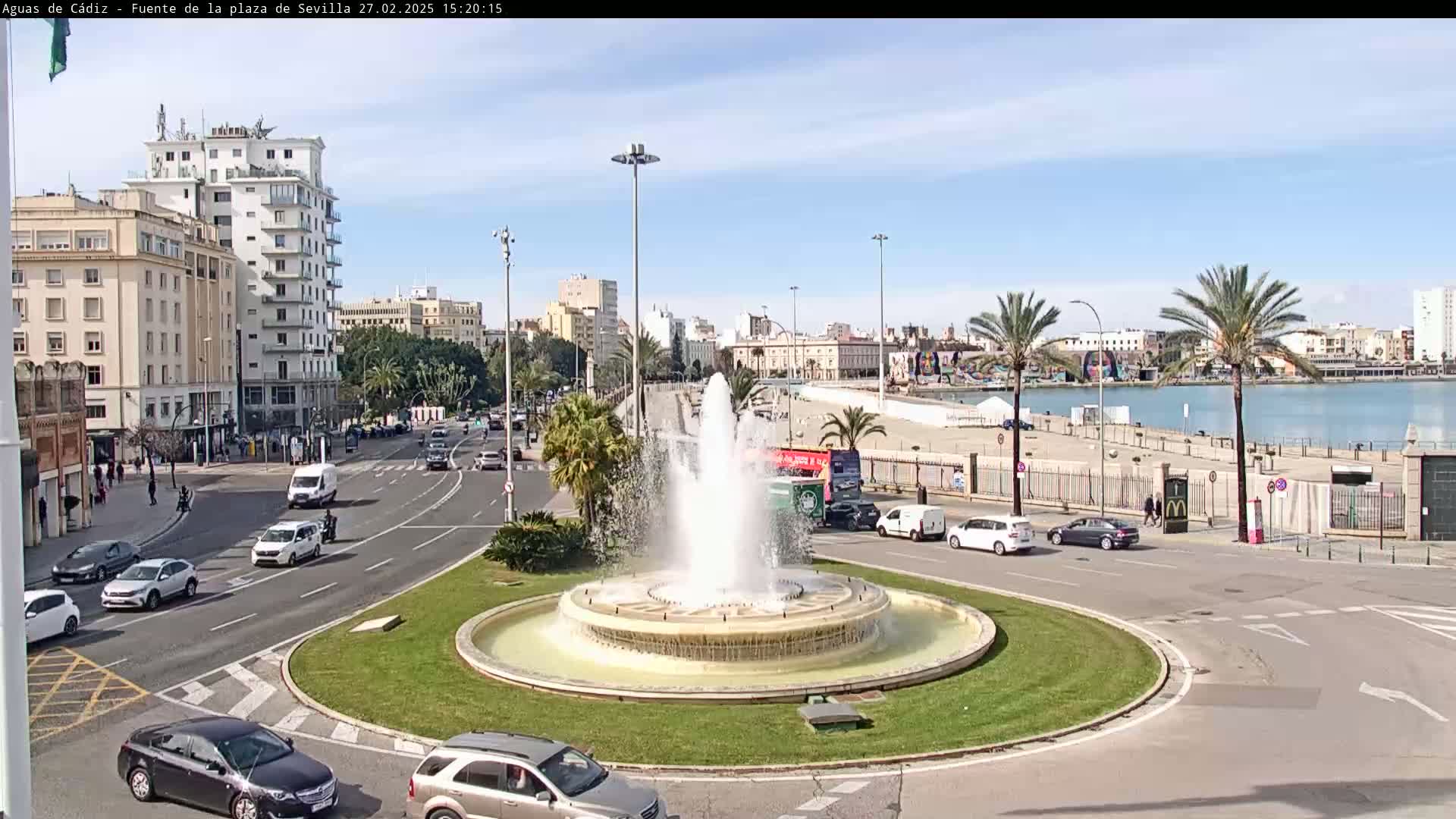 A large fountain sits in a traffic circle, surrounded by city streets, buildings, and palm trees, under a sunny sky near a body of water.