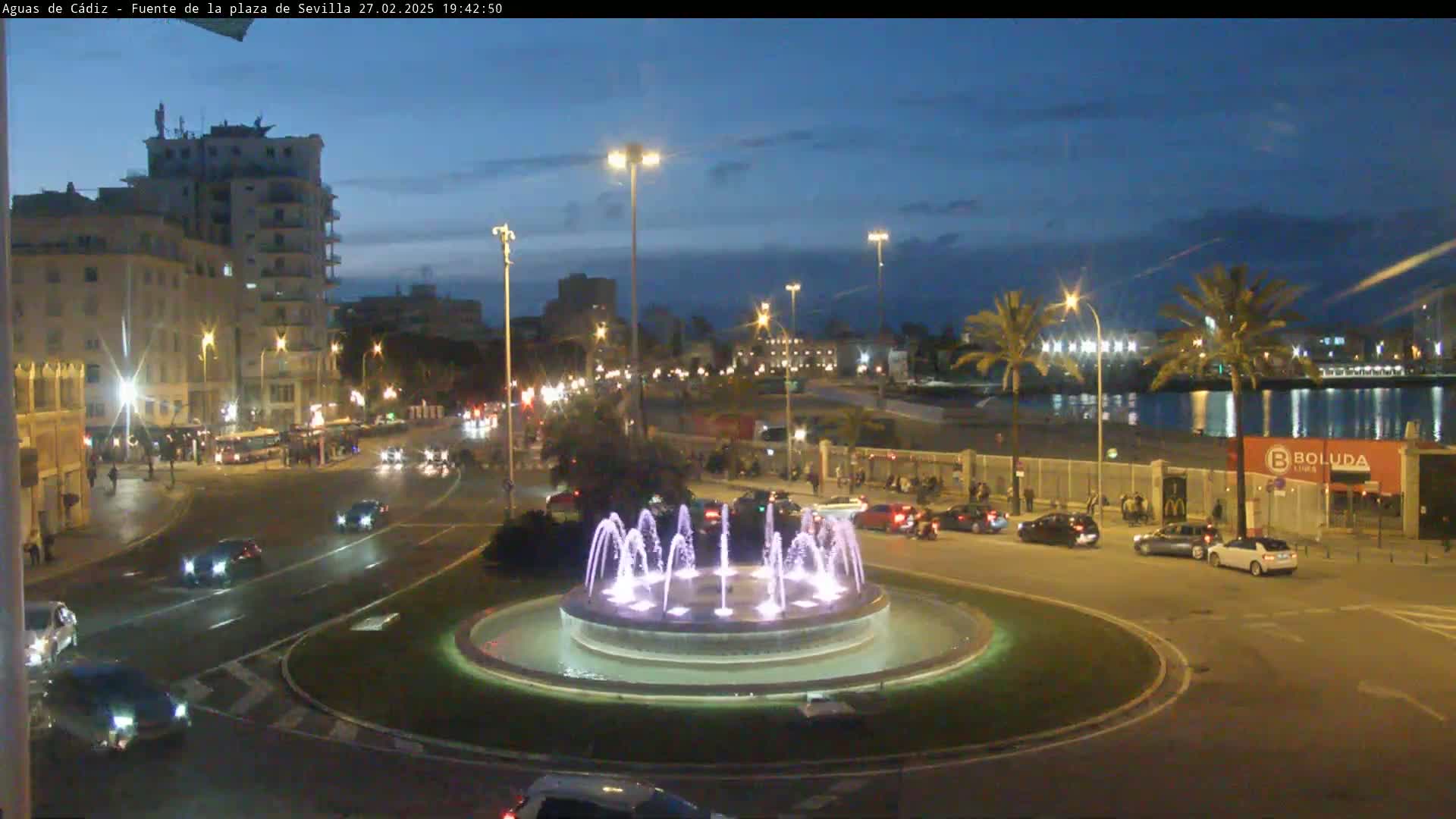 A city roundabout at night, featuring a lit fountain at its center, is surrounded by traffic and buildings under a twilight sky.