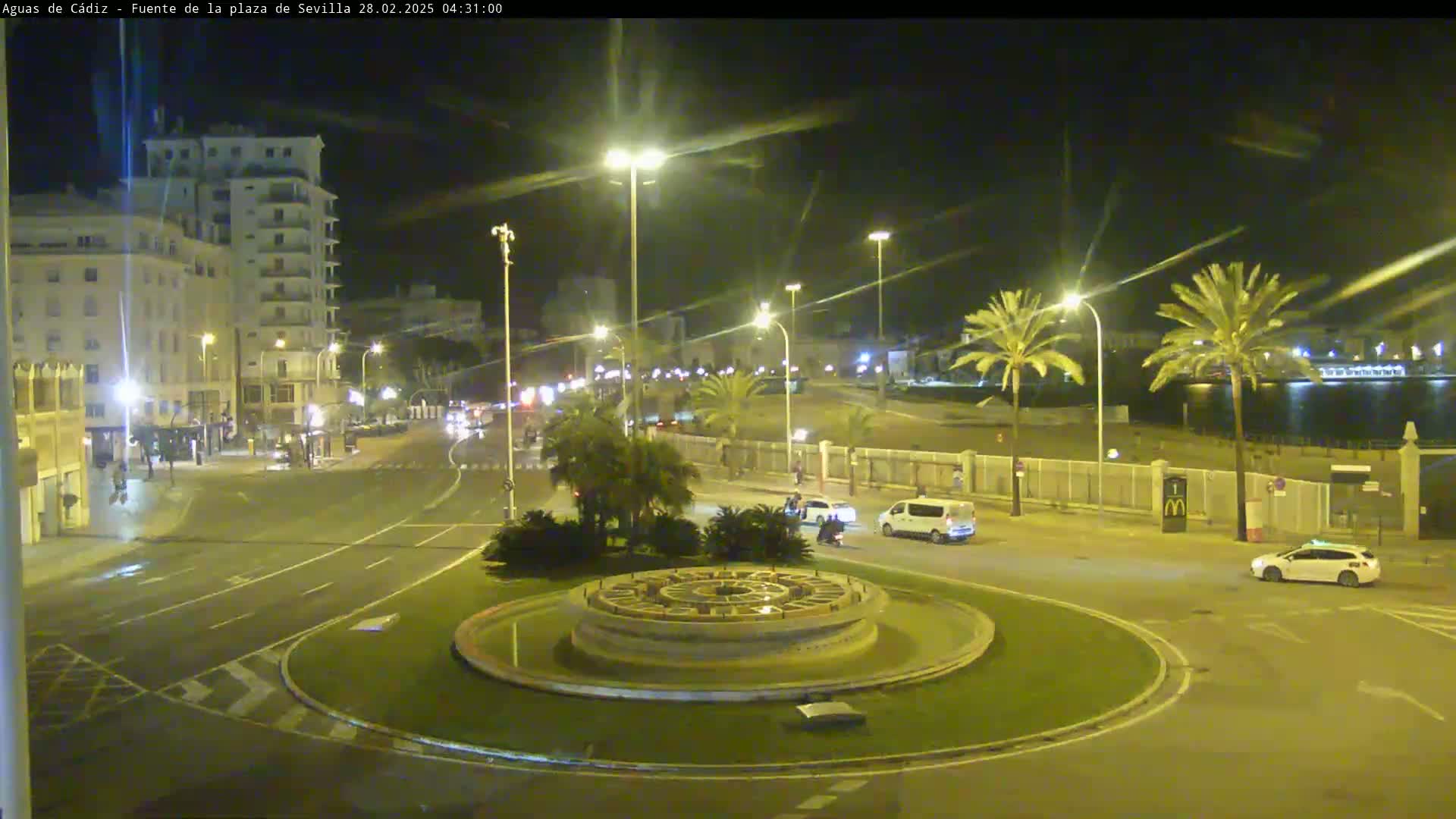 A nighttime view of a city roundabout with a circular fountain at its center, illuminated by streetlights, with palm trees and buildings visible in the background.