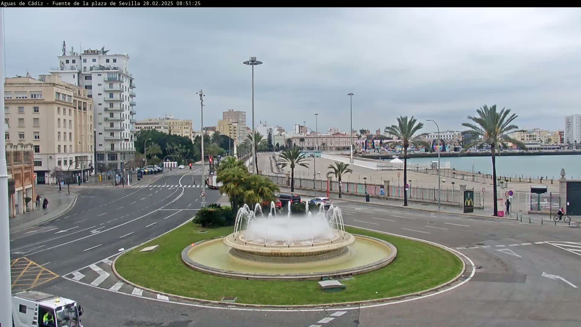 A circular fountain sprays water in a city square on an overcast day, with palm trees and buildings visible in the background, near a body of water.