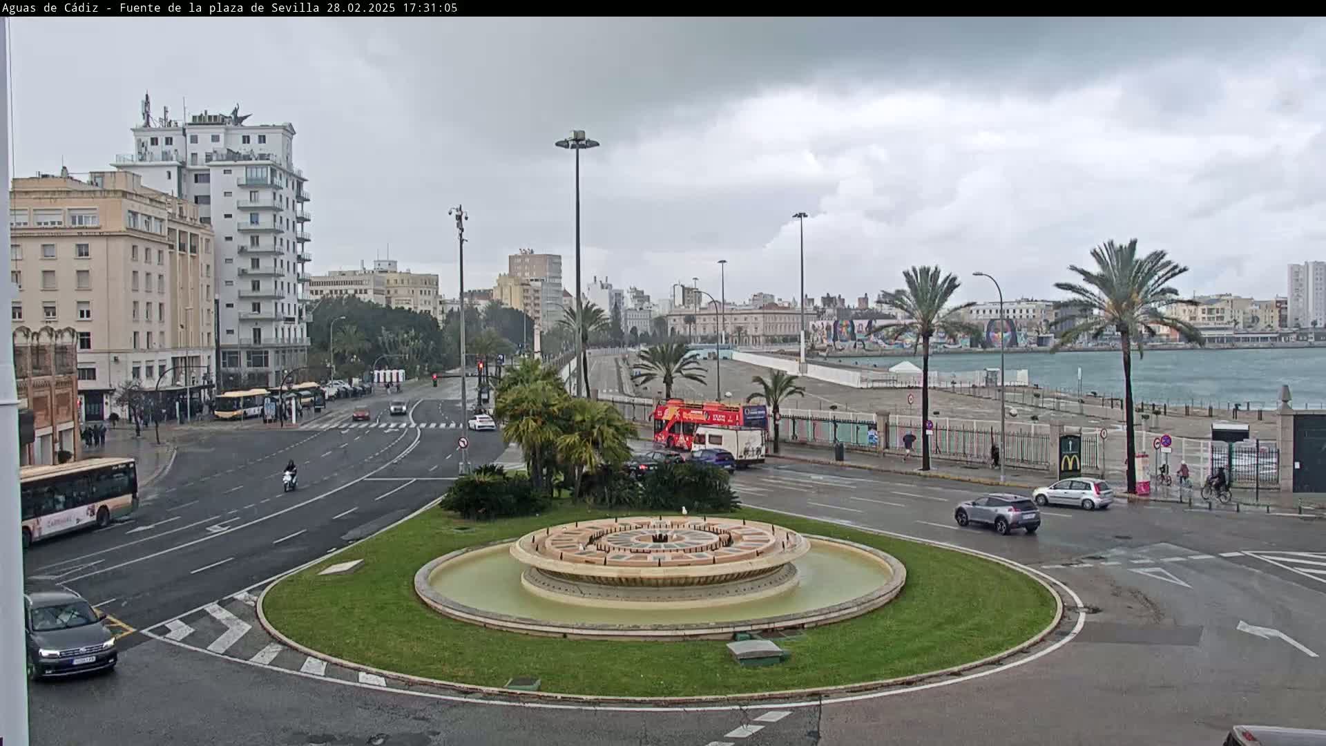 A city roundabout with a fountain in the center, surrounded by roads with cars and buses, under an overcast sky.
