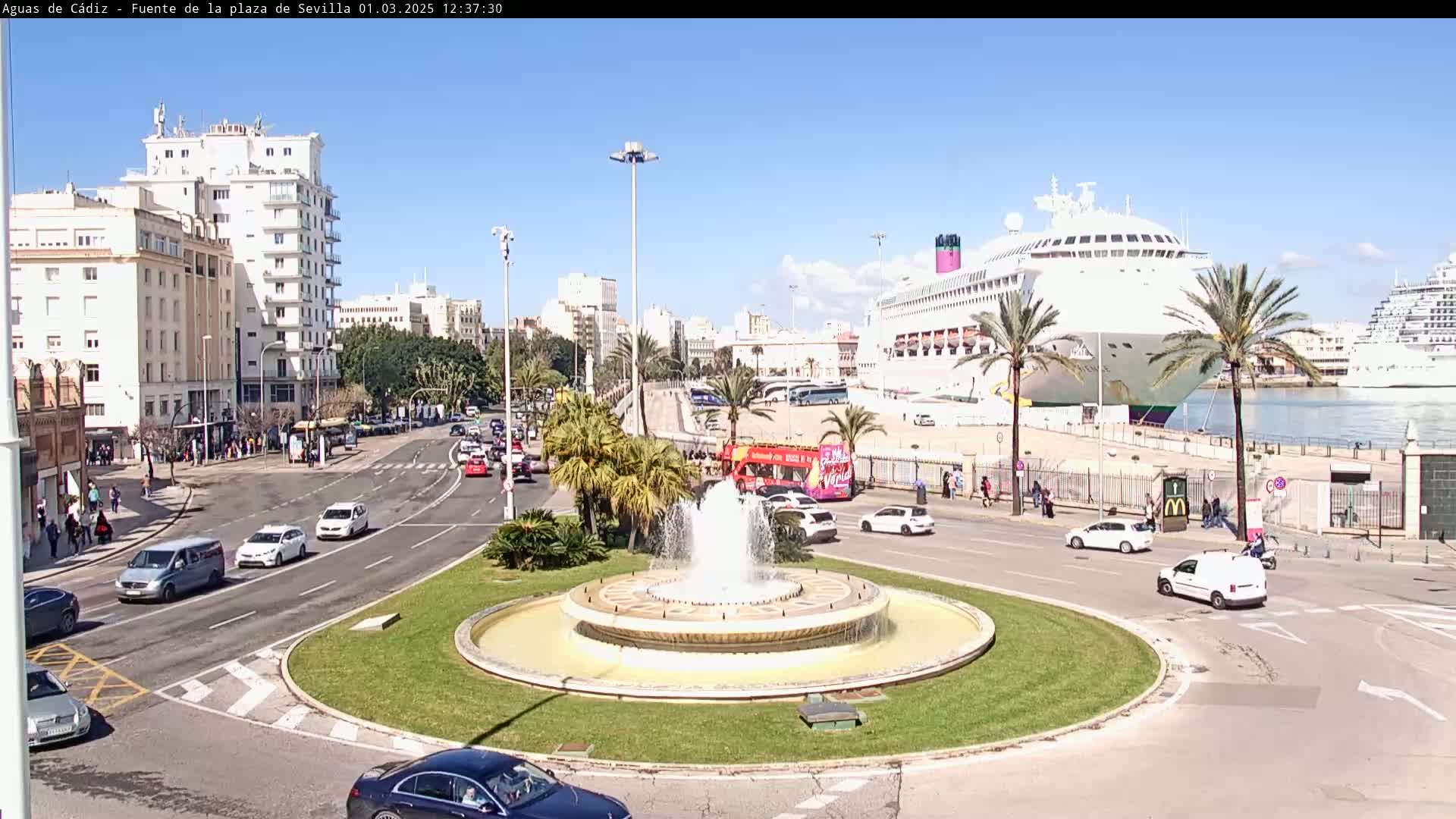 A city street scene on a sunny day features a roundabout with a fountain, several cars, a double-decker bus, and a large cruise ship docked in the harbor.