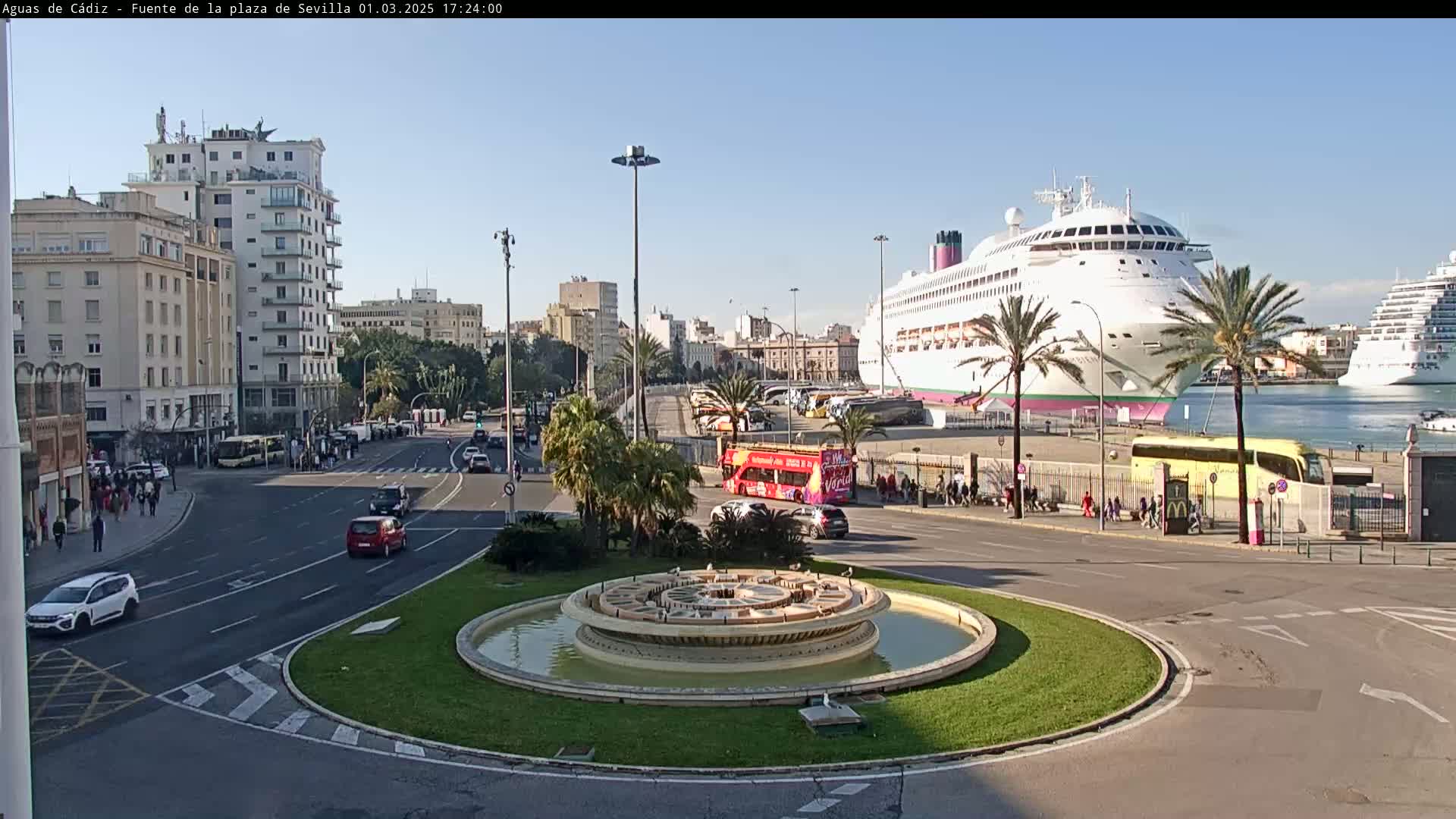 A city roundabout with a circular fountain in the center, overlooks a harbor where a large cruise ship is docked under a clear, sunny sky.