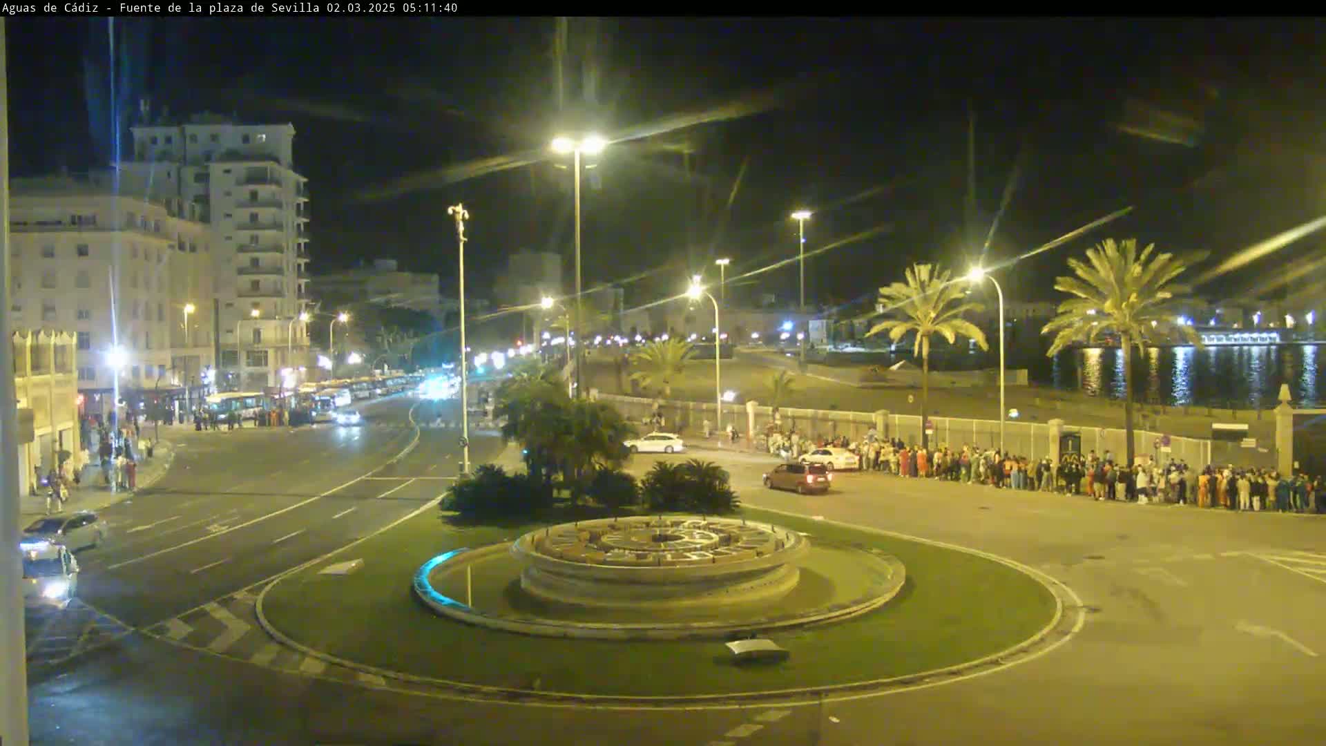 A nighttime, illuminated view of a city roundabout with a circular fountain at its center, featuring vehicles on the road, buildings in the background, and a group of people standing along the curb.