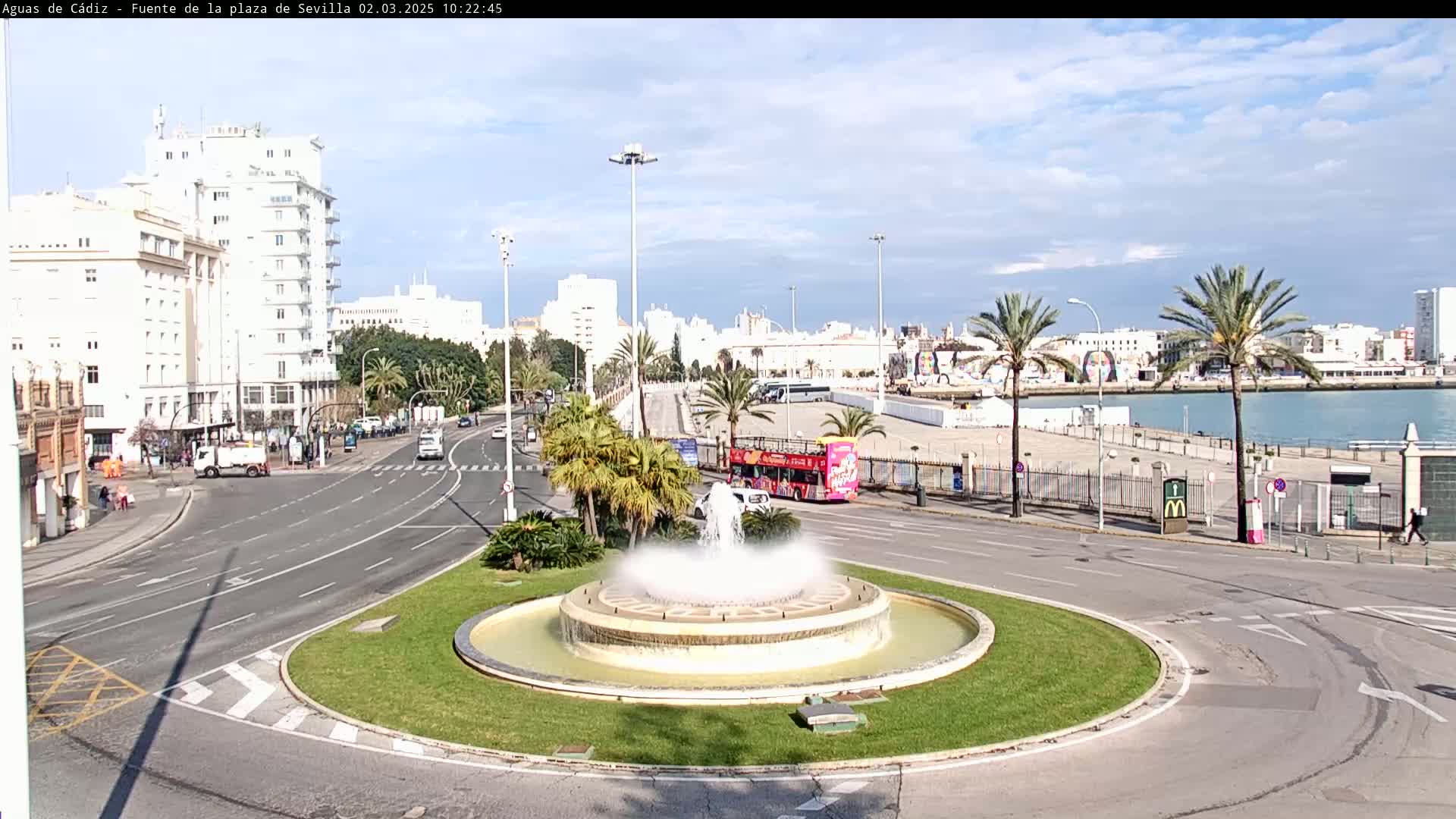 A roundabout with a central fountain is surrounded by roads, buildings, palm trees, and a waterfront under partly cloudy skies.
