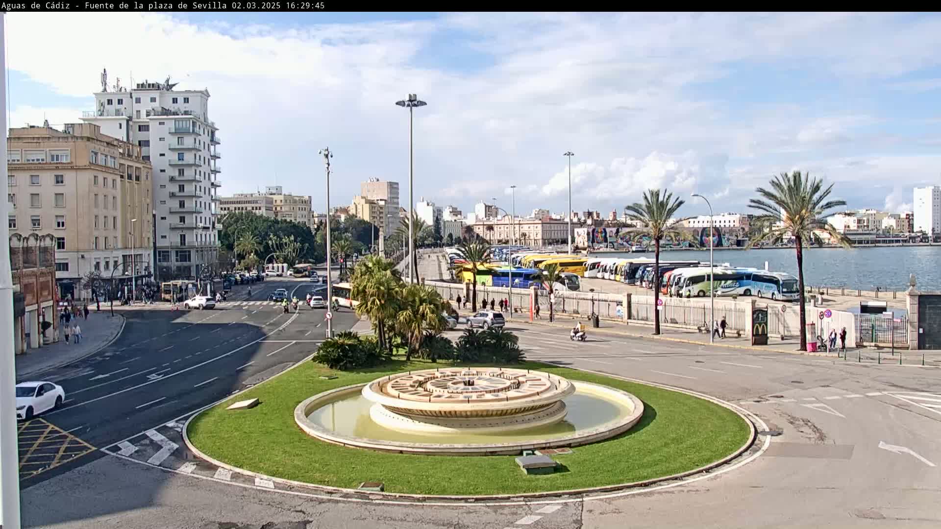 A circular fountain sits in the center of a plaza, surrounded by roadways and buildings, under a partly cloudy sky, with a waterfront lined with numerous buses visible in the background.