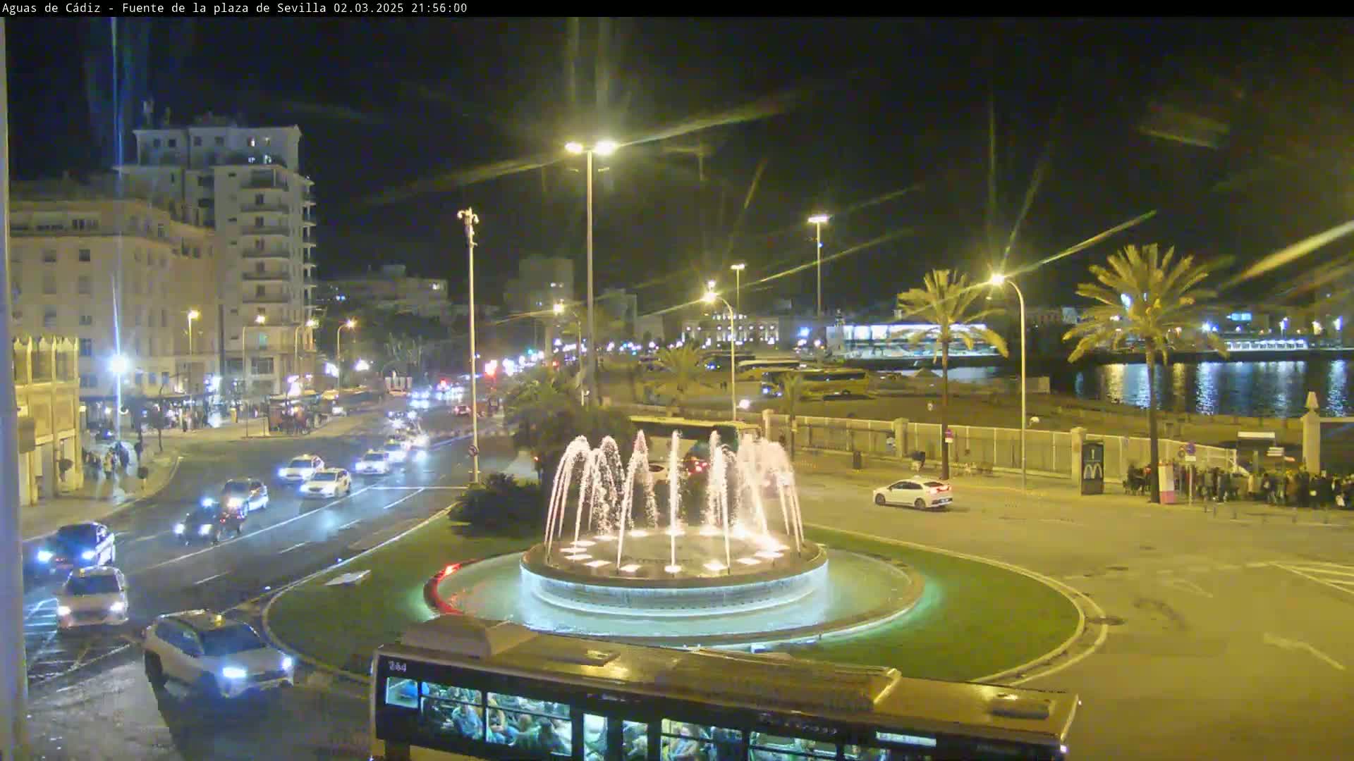 A nighttime view of a city square with a circular fountain, illuminated buildings, and traffic on surrounding roads, under clear skies.
