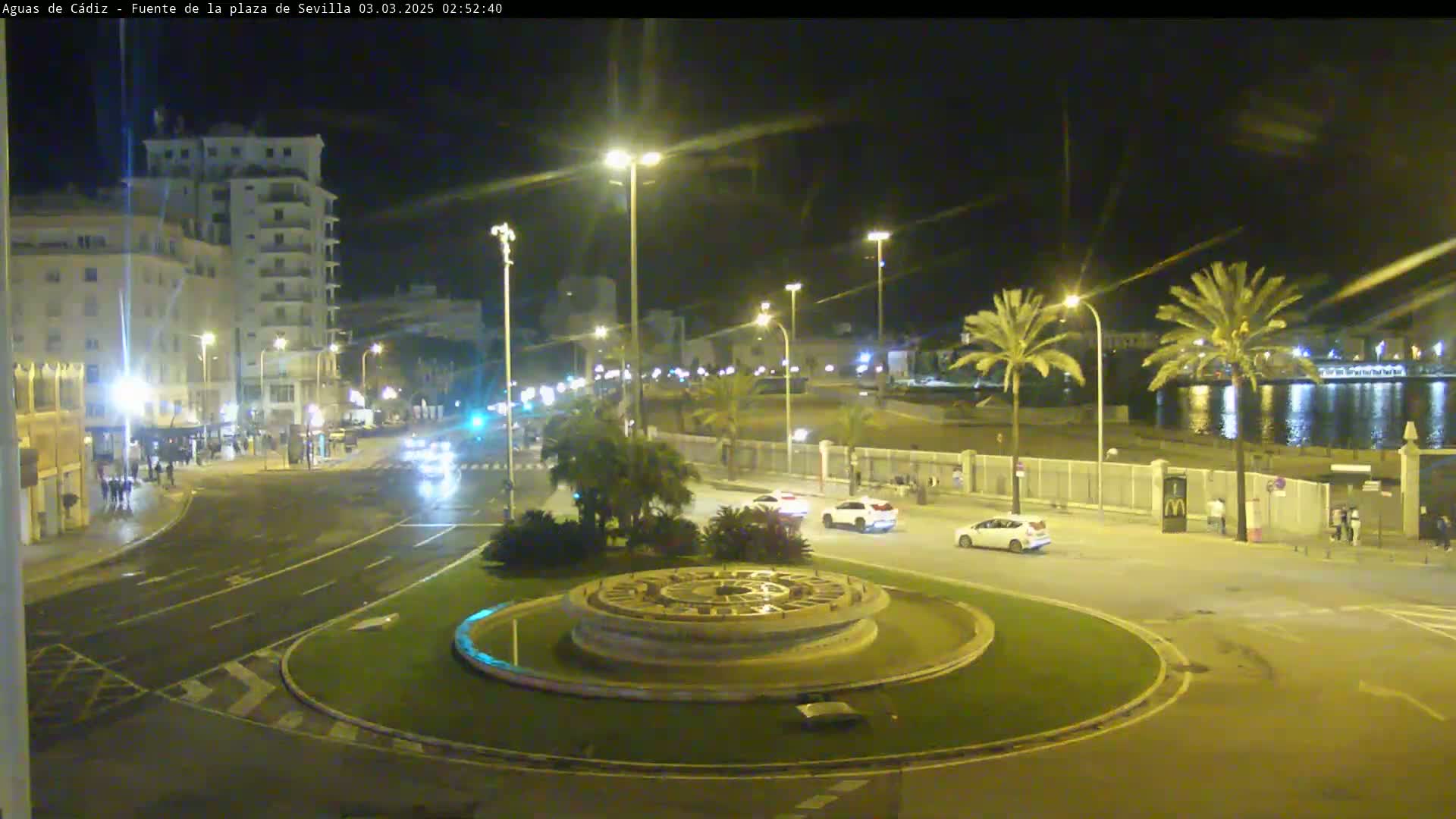 A nighttime view of a city roundabout with a circular fountain at its center, several cars, and palm trees lining a waterfront in the background.
