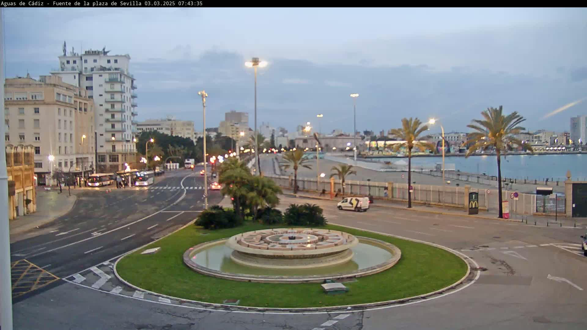 A circular fountain sits in a traffic circle near a waterfront, with buildings and palm trees lining the streets under a partly cloudy sky.