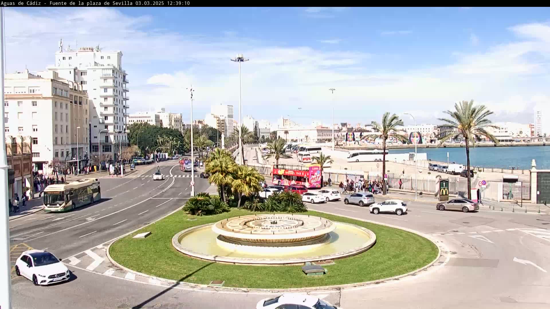 A circular fountain sits in the center of a traffic circle, surrounded by palm trees, buildings, and various vehicles under a mostly clear sky.