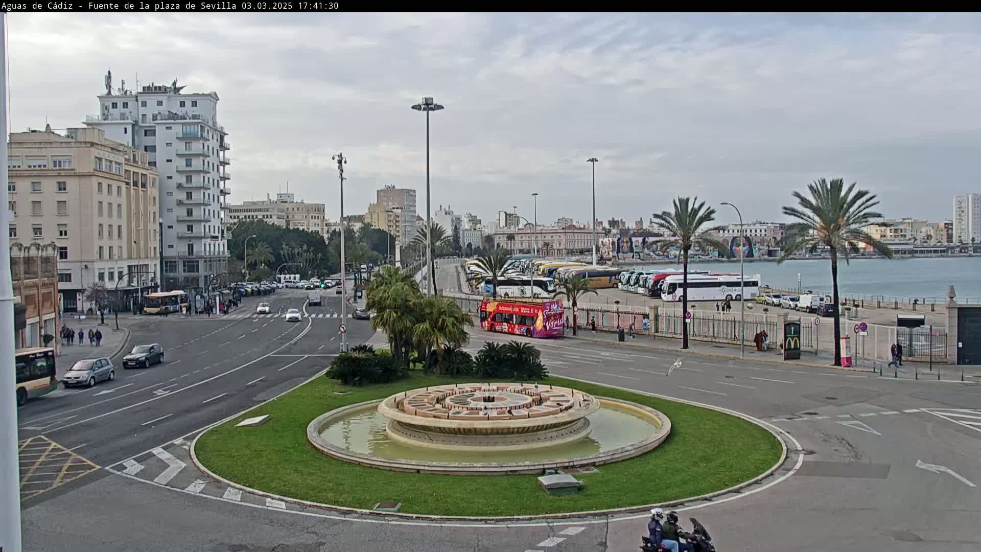 A city square with a circular fountain in the center, surrounded by roads, buildings, palm trees, and parked buses, under an overcast sky.