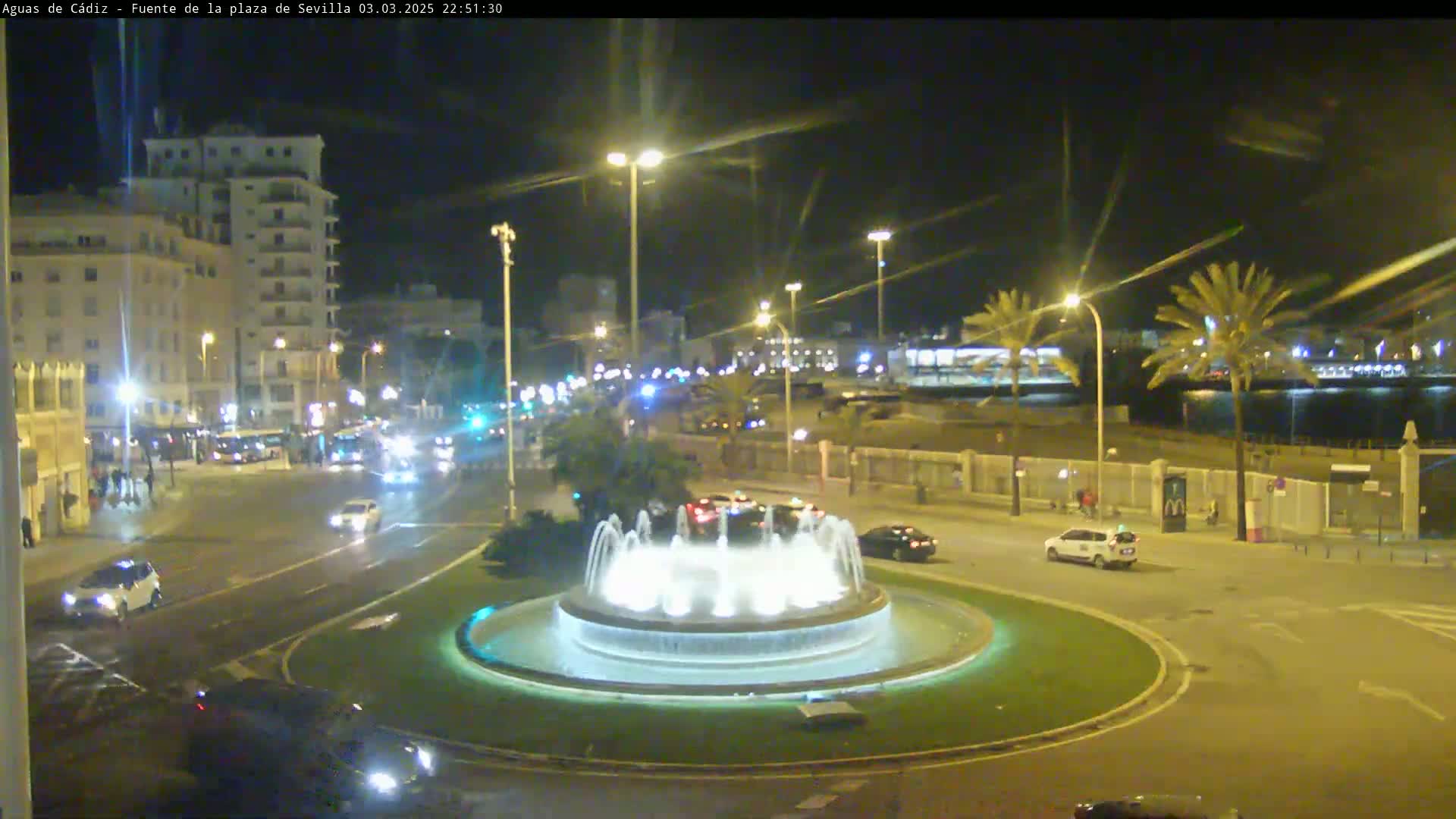 A nighttime view of a city roundabout featuring a lit fountain, with cars driving around it and buildings visible in the background under a clear sky.
