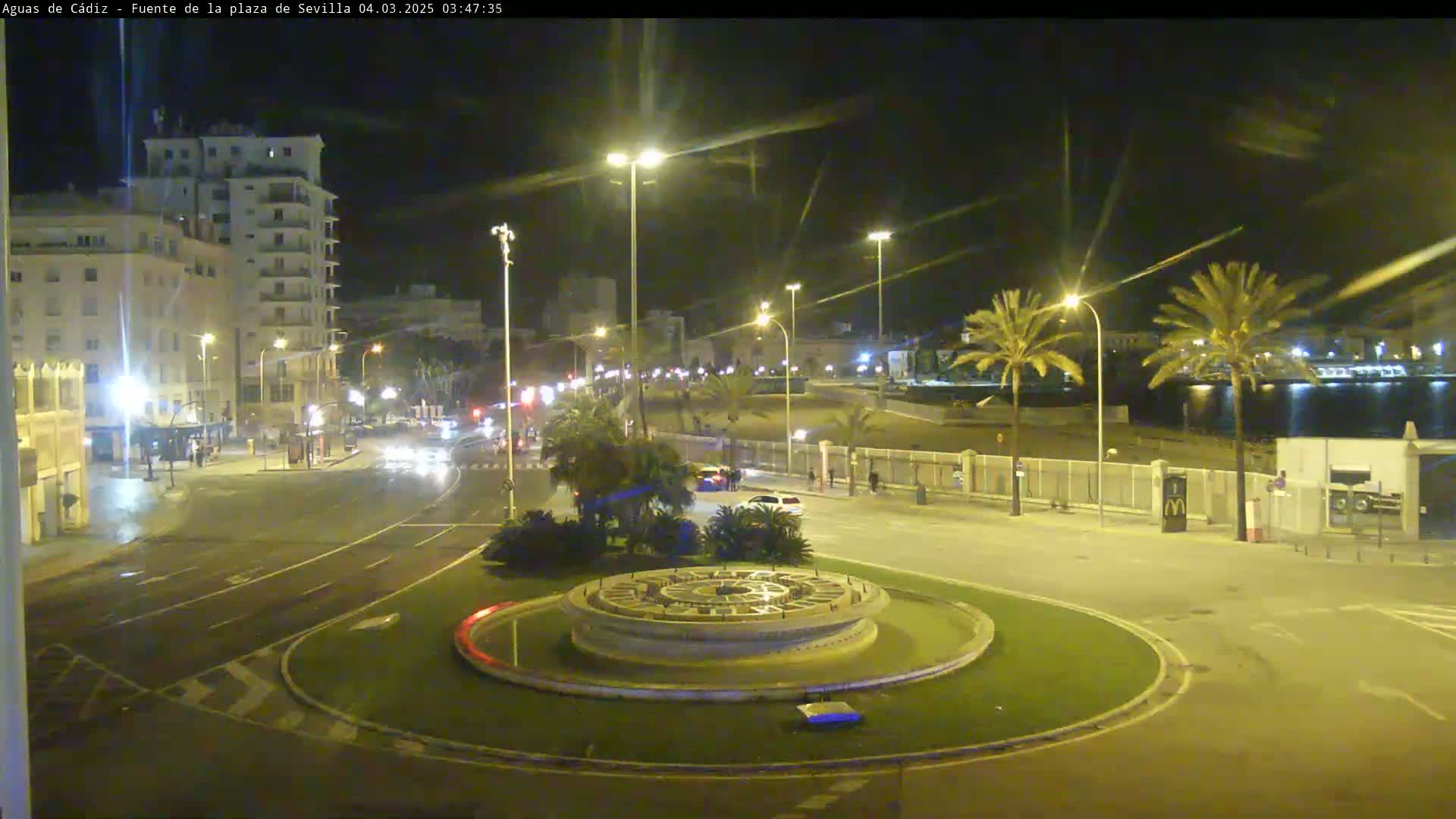 A nighttime view of a circular traffic island with a fountain in the center, surrounded by palm trees, buildings, and a roadway with sparse traffic.