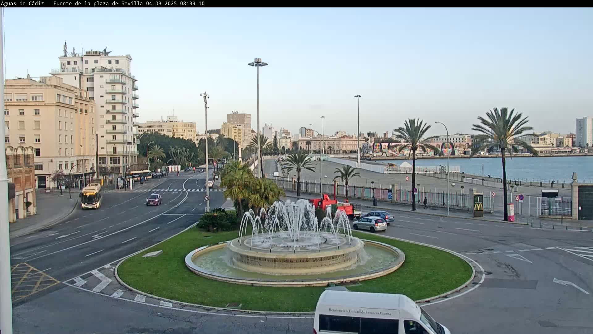 A city street scene with a fountain in a traffic circle, several buildings, palm trees, and a body of water in the background under a clear, bright sky.