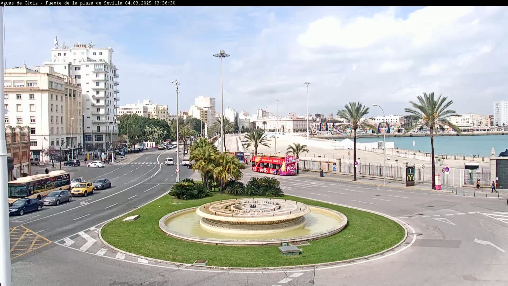 A city roundabout with a circular fountain in the center, surrounded by palm trees, buildings, and vehicles, on a partly cloudy day with a body of water visible in the background.