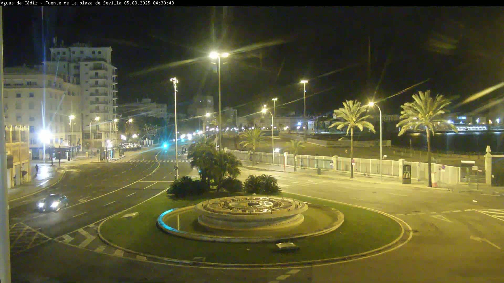 A nighttime, brightly lit roundabout with a circular fountain at its center, surrounded by roads, buildings, and palm trees.
