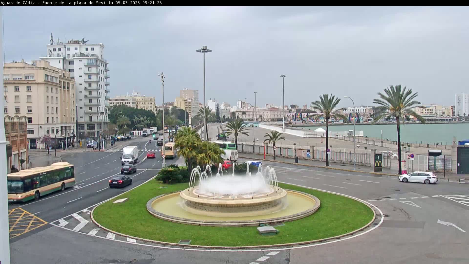 A city roundabout with a fountain in the center, surrounded by buildings, palm trees, and several cars and buses, on an overcast day.