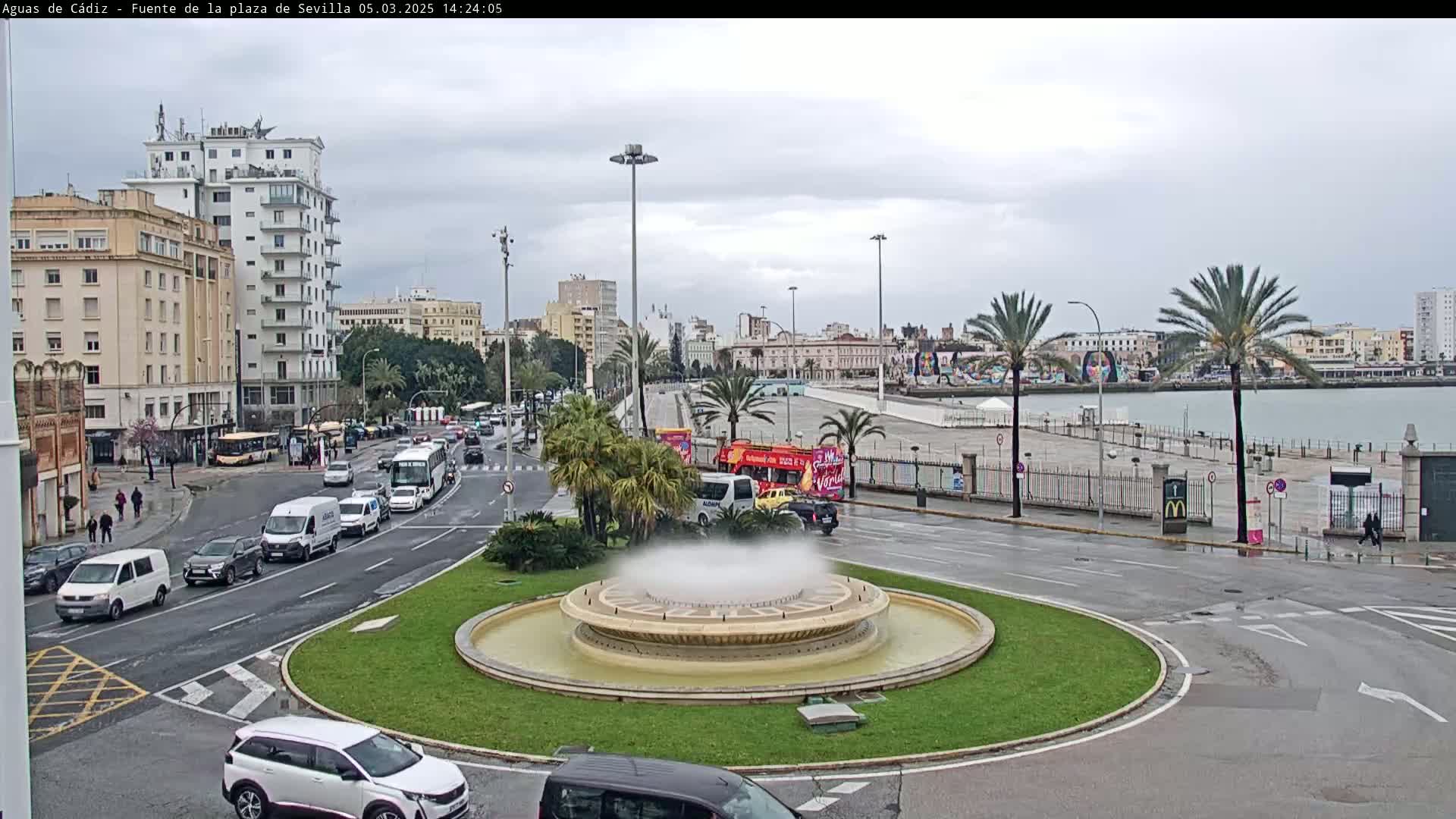 A city roundabout with a fountain in the center, surrounded by traffic and buildings, on an overcast day near a waterfront.