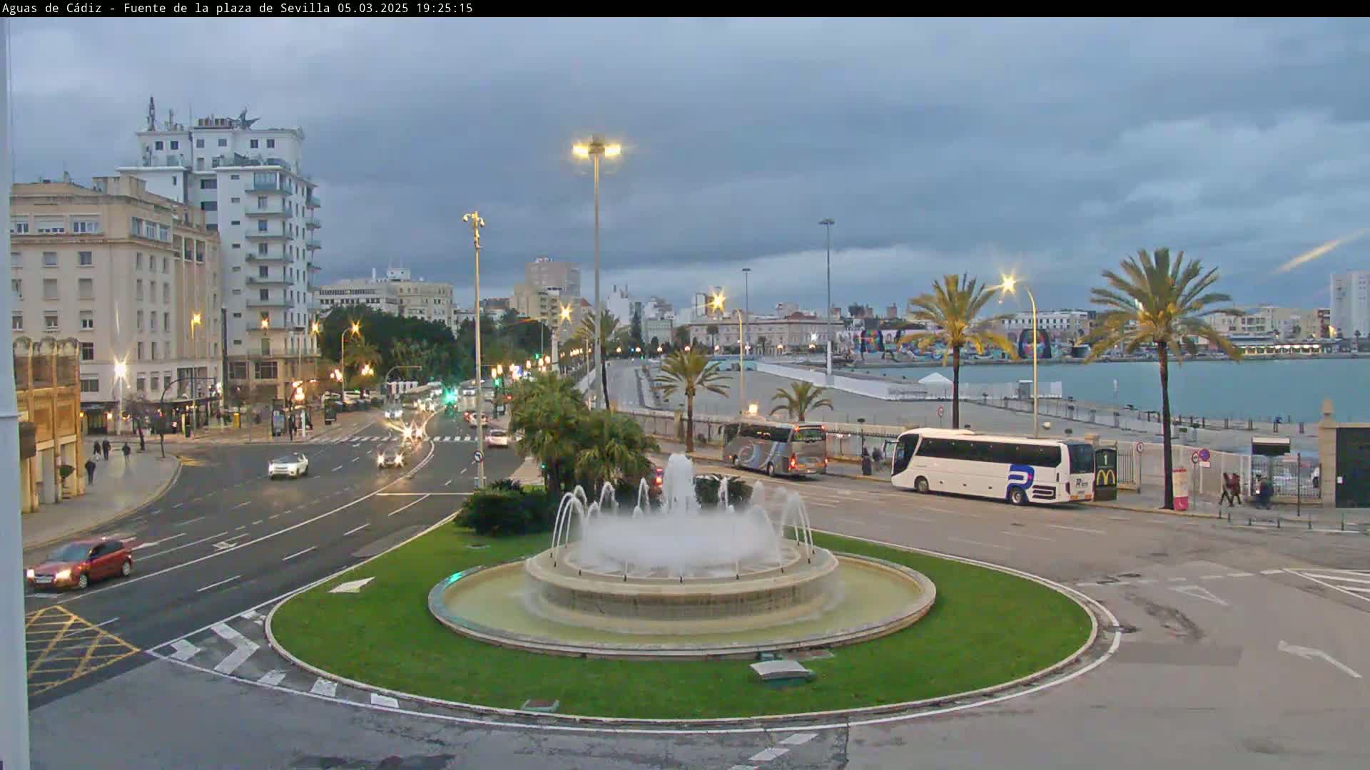 A city fountain in a traffic circle is surrounded by buildings, palm trees, and cars under an overcast sky.