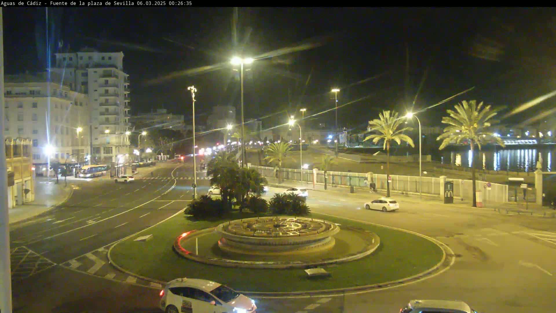 A nighttime view of a city roundabout with a fountain at its center, surrounded by palm trees, buildings, and cars driving on the roadway next to a calm body of water.