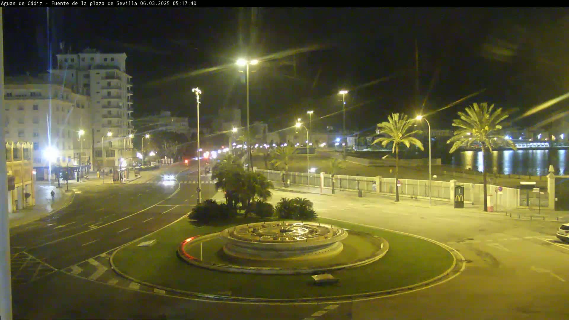 A nighttime view of a city roundabout with a fountain at its center, surrounded by palm trees, buildings, and a calm body of water in the background.