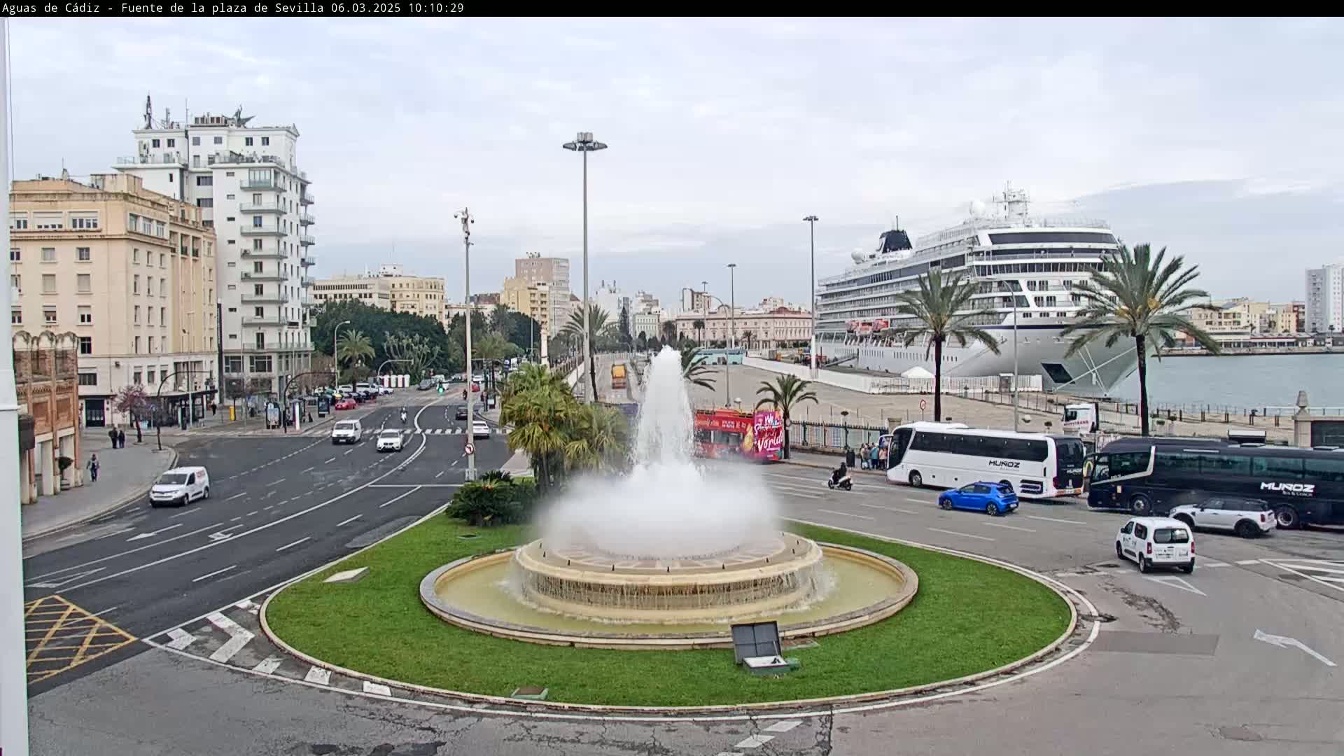 A large fountain is in the center of a roundabout in a city, with a cruise ship docked in the harbor in the background on an overcast day.