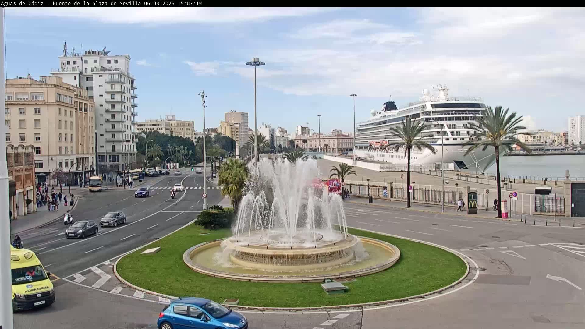 A large cruise ship is docked at a port, visible from a city roundabout featuring a central fountain, with light traffic and partly sunny weather conditions.