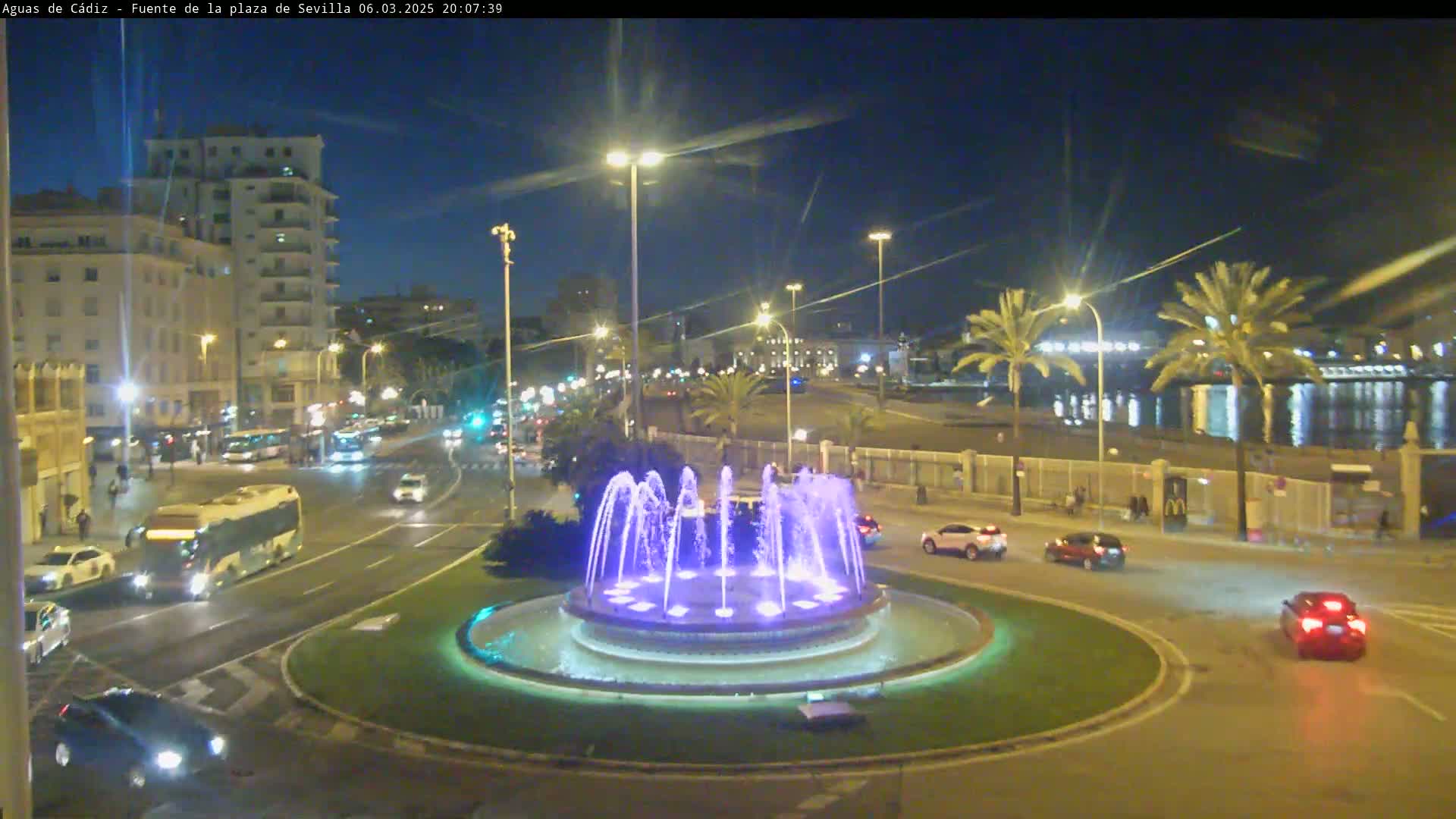 A nighttime view of a city roundabout featuring a circular fountain with illuminated water jets, surrounded by streets with cars and a bus, and buildings and palm trees in the background under a clear, dark sky.