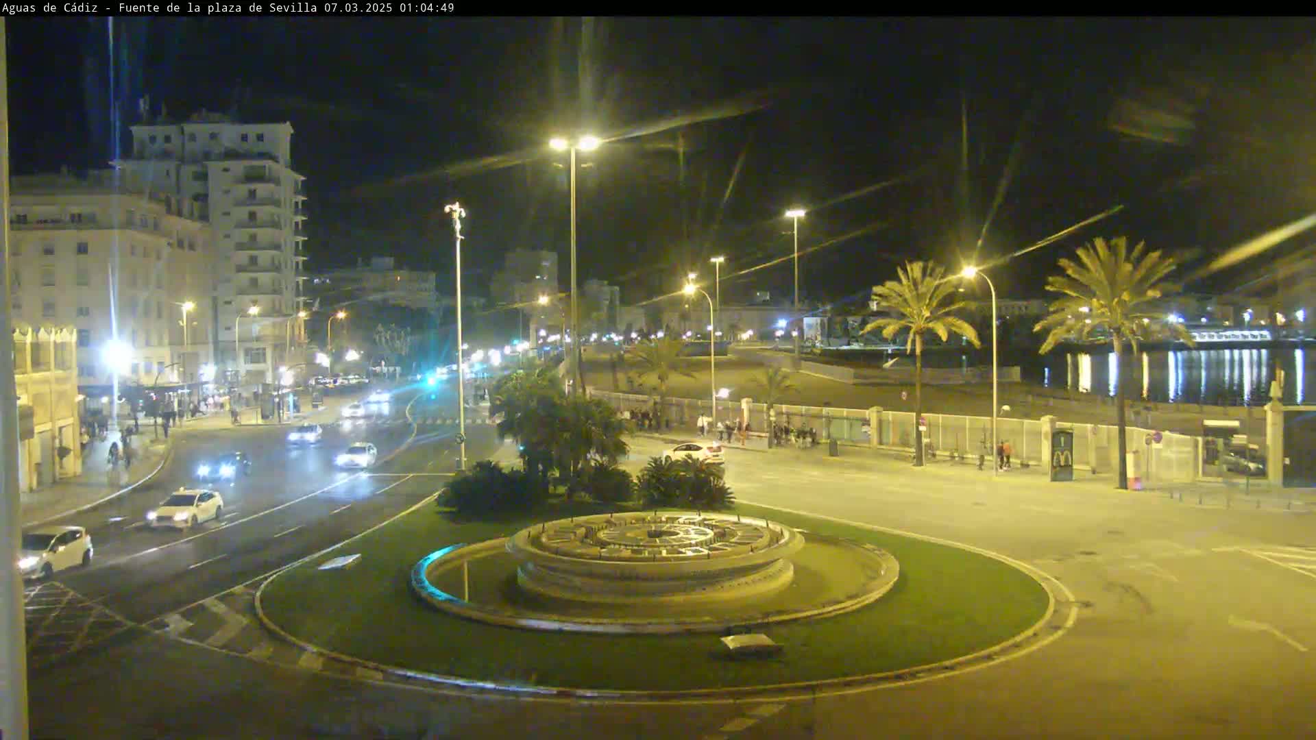 A nighttime street scene shows a roundabout with a fountain in the center, surrounded by palm trees, buildings, and cars driving on a wet roadway under bright streetlights.