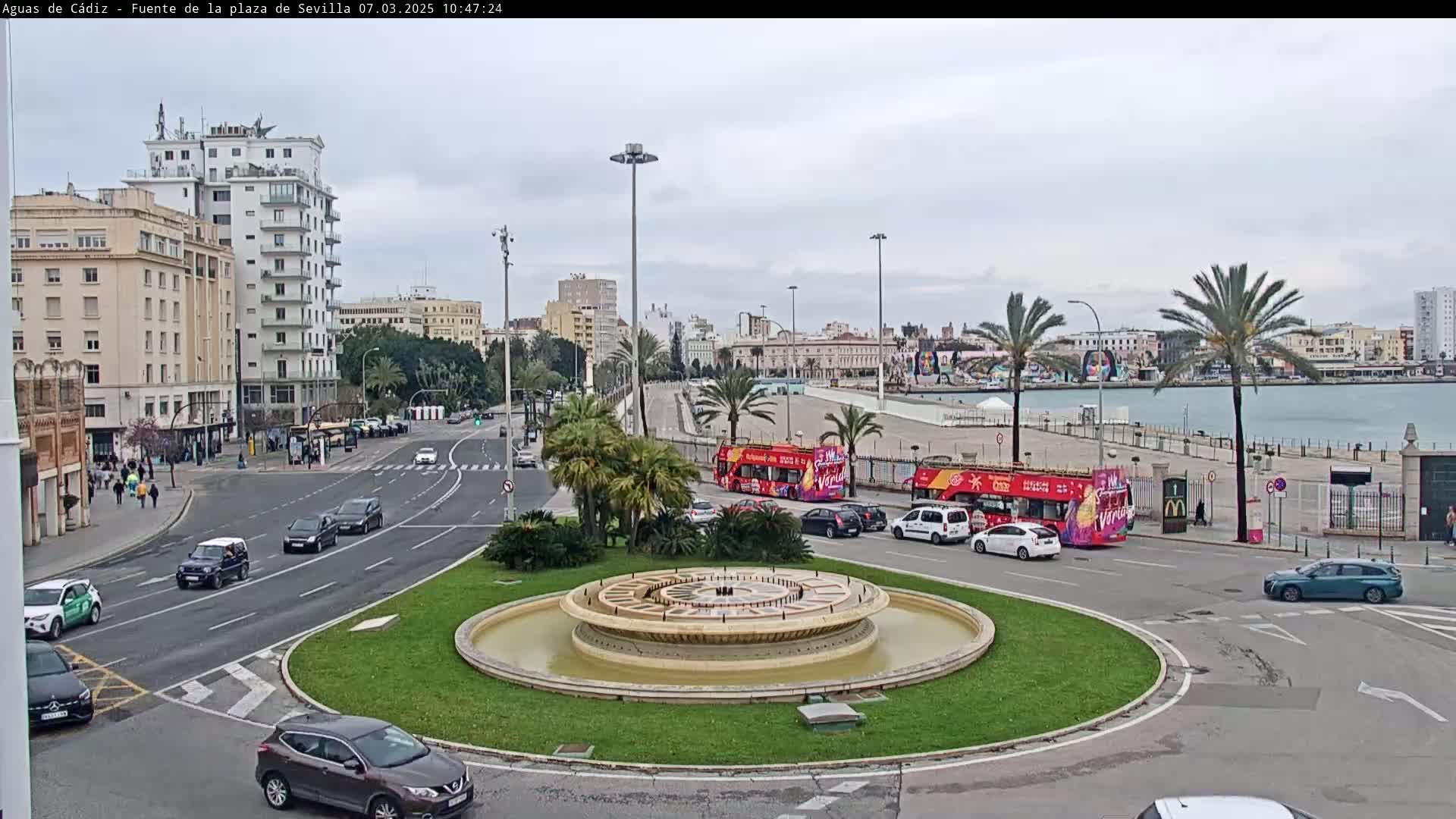 A city square with a circular fountain in the center, surrounded by streets with cars and buildings, on an overcast day near a body of water.
