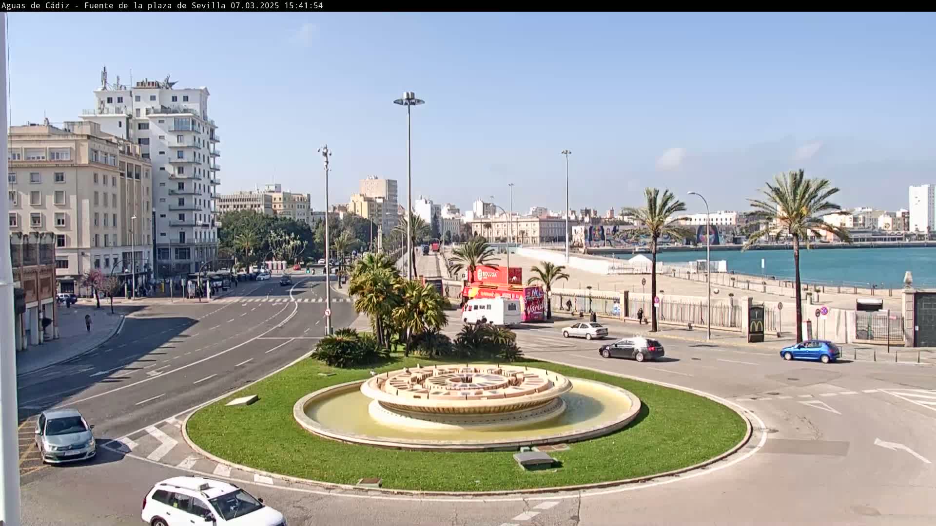 A city roundabout with a central fountain is surrounded by roads, buildings, palm trees, and a waterfront with calm blue water under a clear, sunny sky.