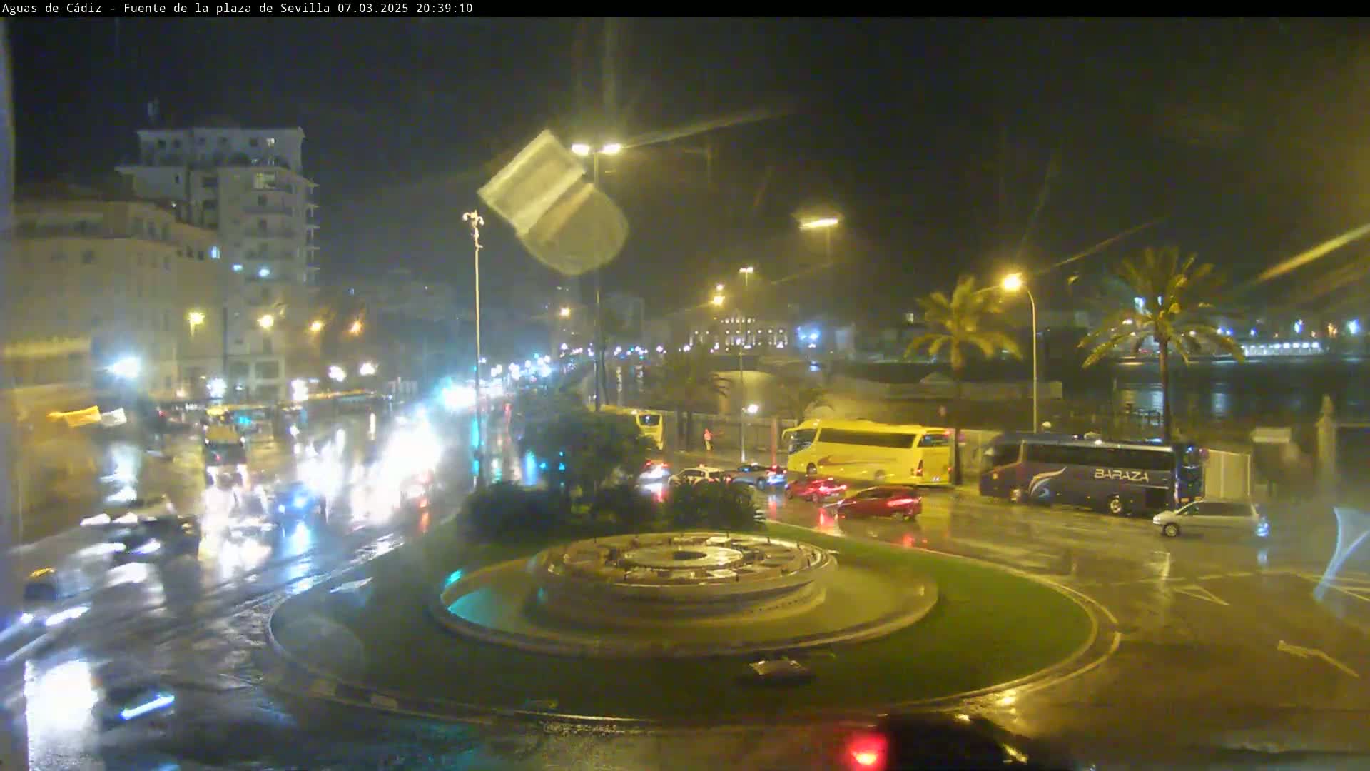 A nighttime scene of a rain-slicked roundabout with traffic, buildings, palm trees, and a circular fountain in the center.