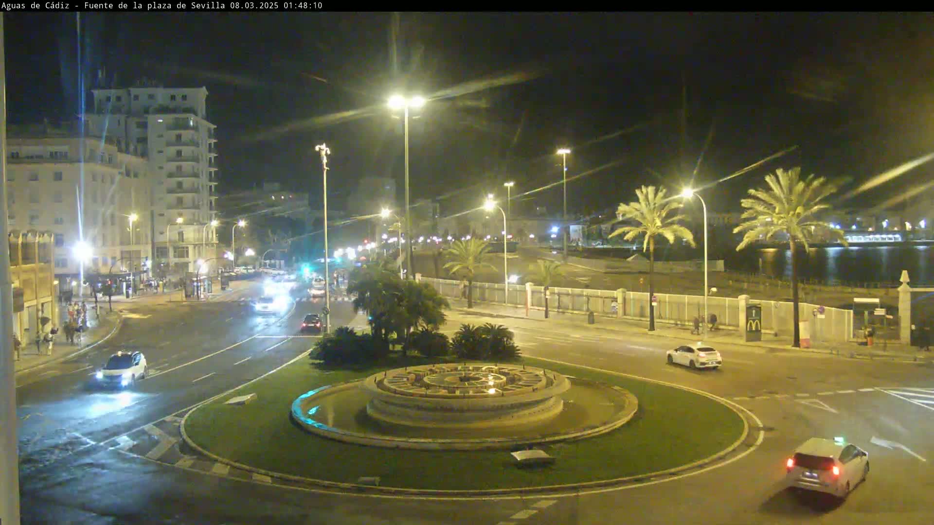 A nighttime view of a roundabout with a fountain in the center, surrounded by streets with cars and buildings, under illuminated streetlights.