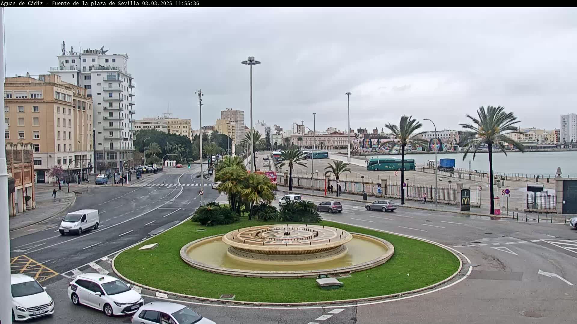 A roundabout with a fountain in the center, surrounded by city streets, buildings, and palm trees on a cloudy day.