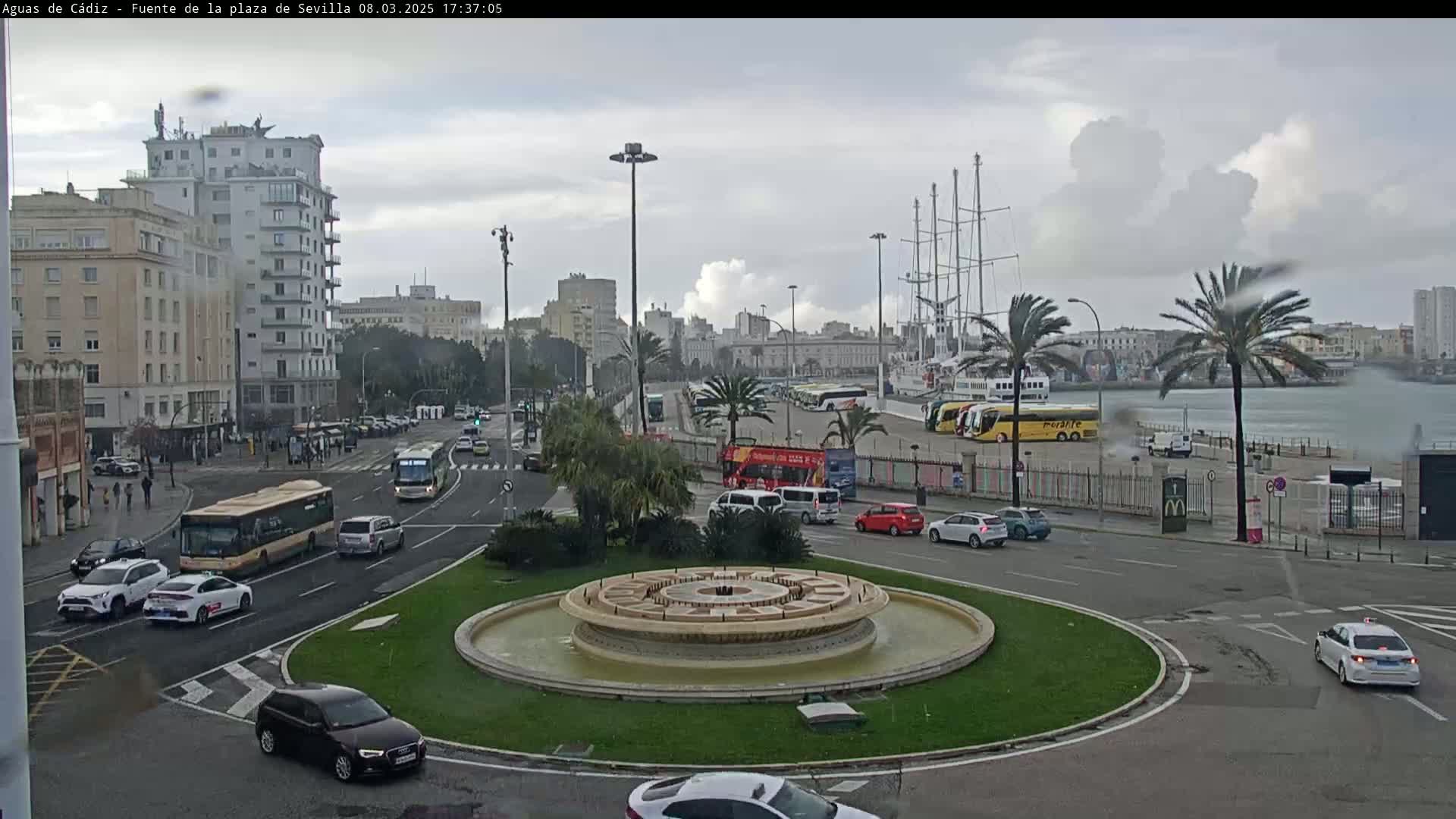 A city roundabout with a fountain in the center, surrounded by cars and buses, overlooks a harbor with docked ships under a cloudy, overcast sky with light rain.