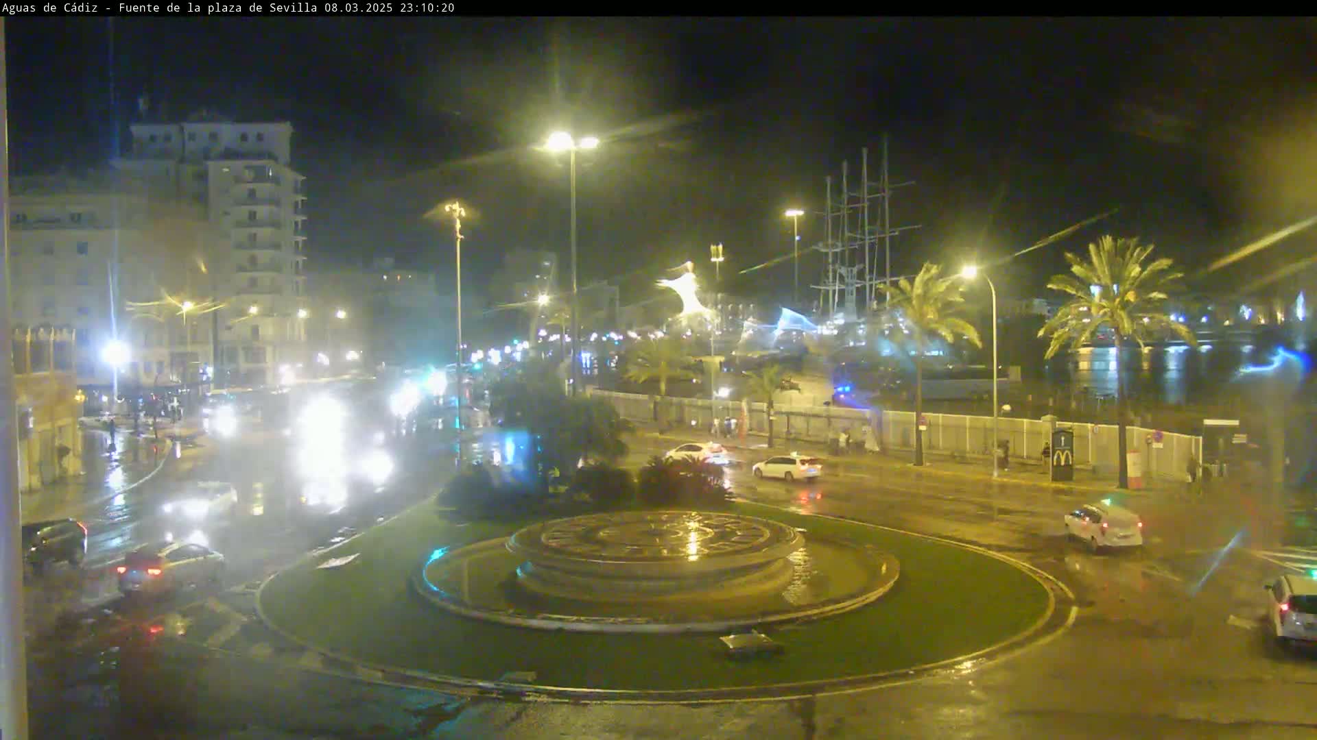 A nighttime, rain-slicked roundabout with a large fountain at its center is surrounded by city streets with cars and buildings under streetlights.