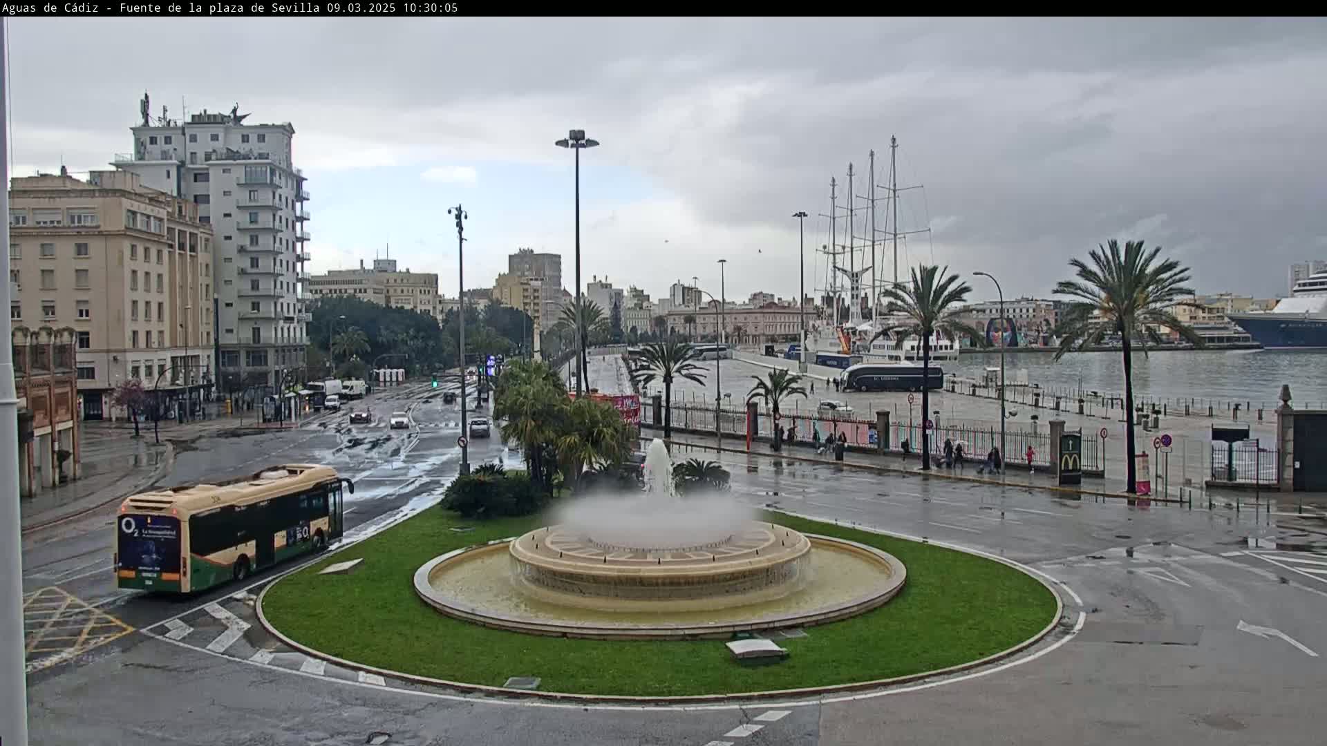 A city roundabout with a fountain in the center is shown on a rainy day, with a harbor and docked ships in the background.