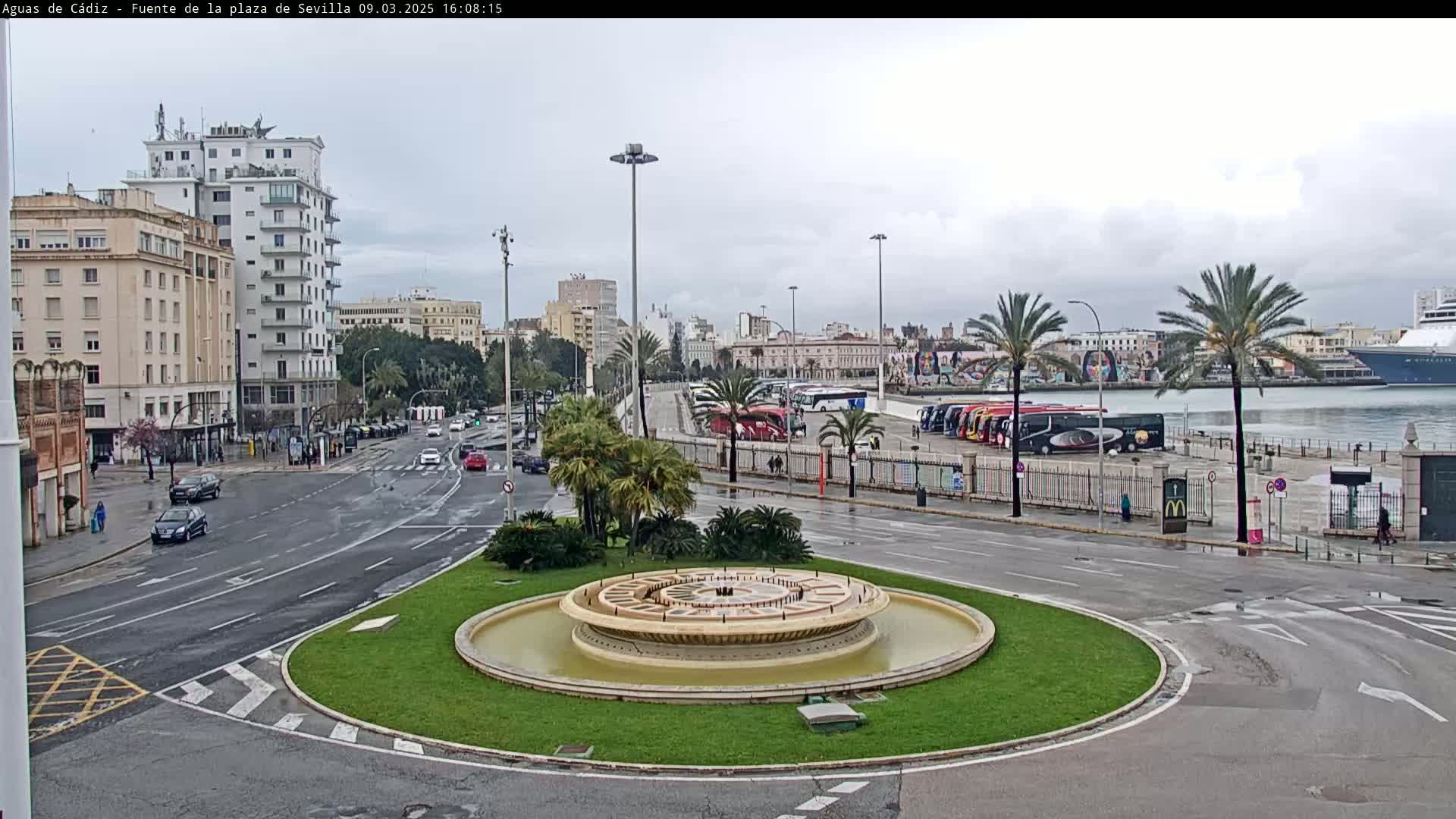 A circular fountain sits in a traffic circle, with city buildings, parked tour buses, and a cruise ship visible in the background on a cloudy day.