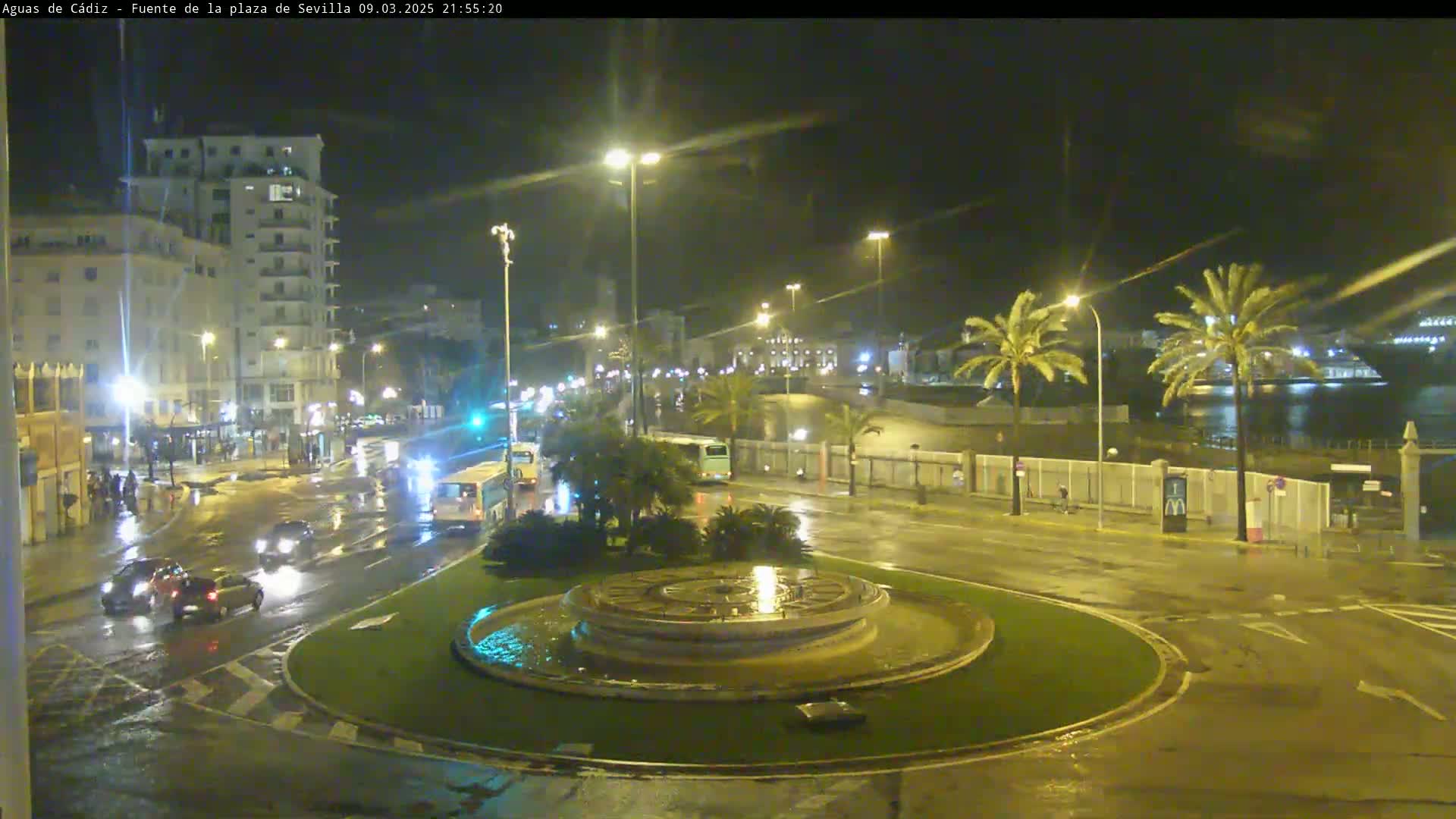 A wet, nighttime scene shows a roundabout with a fountain in the center, surrounded by city streets with cars and buses driving on them, under streetlights.