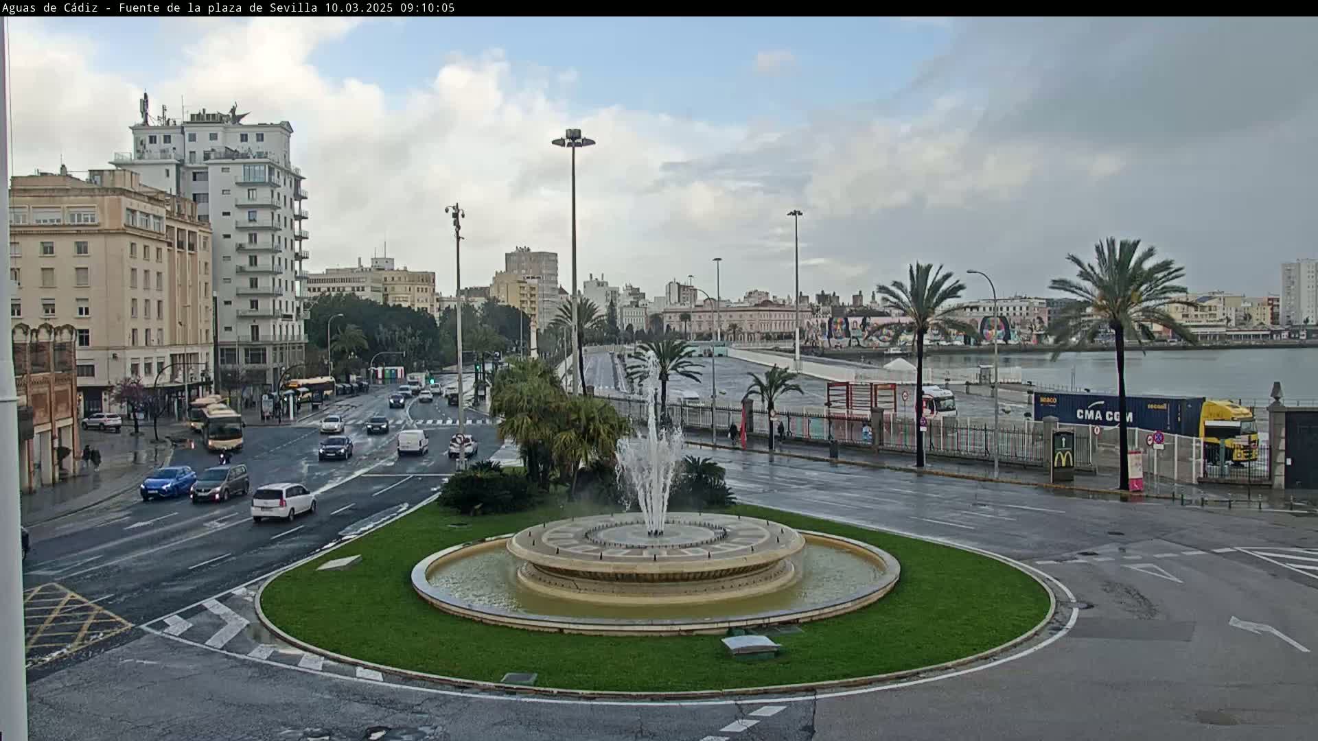 A city roundabout with a fountain in the center, wet streets, and buildings visible in the background under an overcast sky.