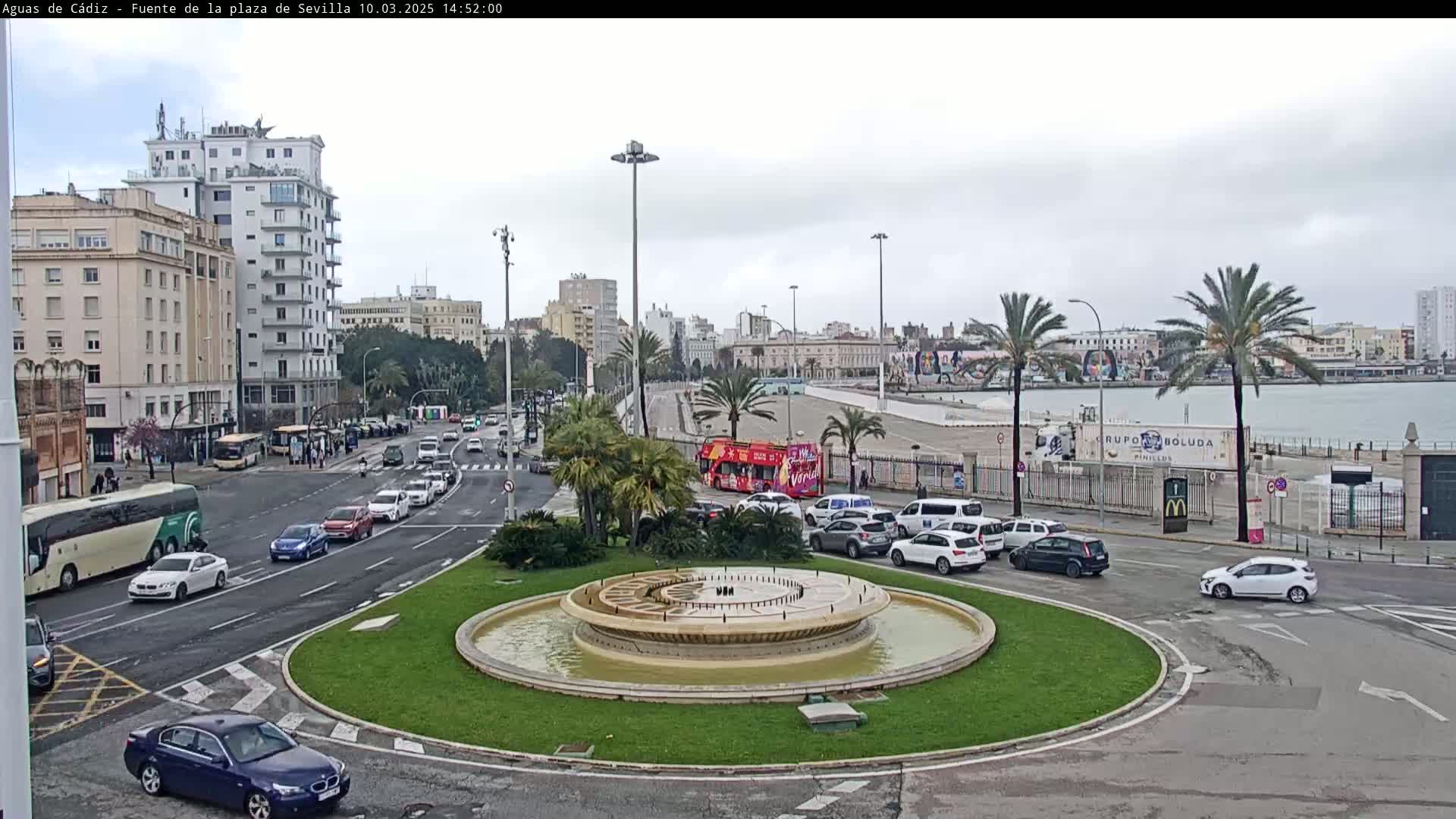 A city roundabout with a circular fountain in the center, surrounded by palm trees, buildings, and various vehicles under an overcast sky.
