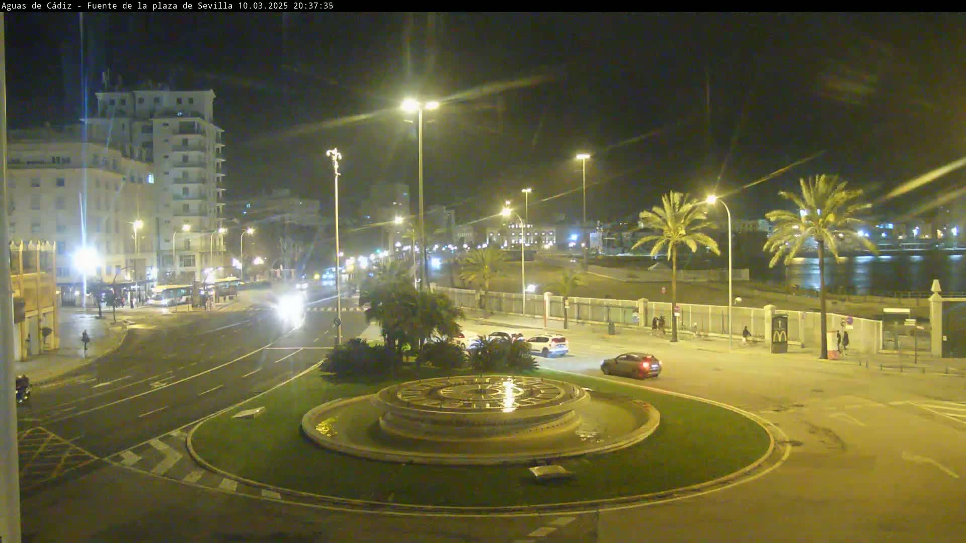 A nighttime view of a city roundabout with a circular fountain, illuminated by streetlights, with cars driving on the roads and palm trees lining a nearby waterfront.