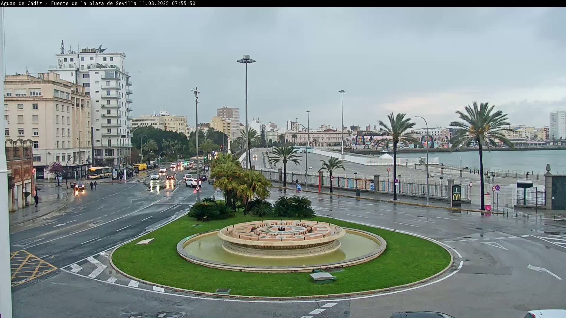 A city roundabout with a circular fountain in the center, surrounded by palm trees, buildings, and wet roads under a cloudy sky.