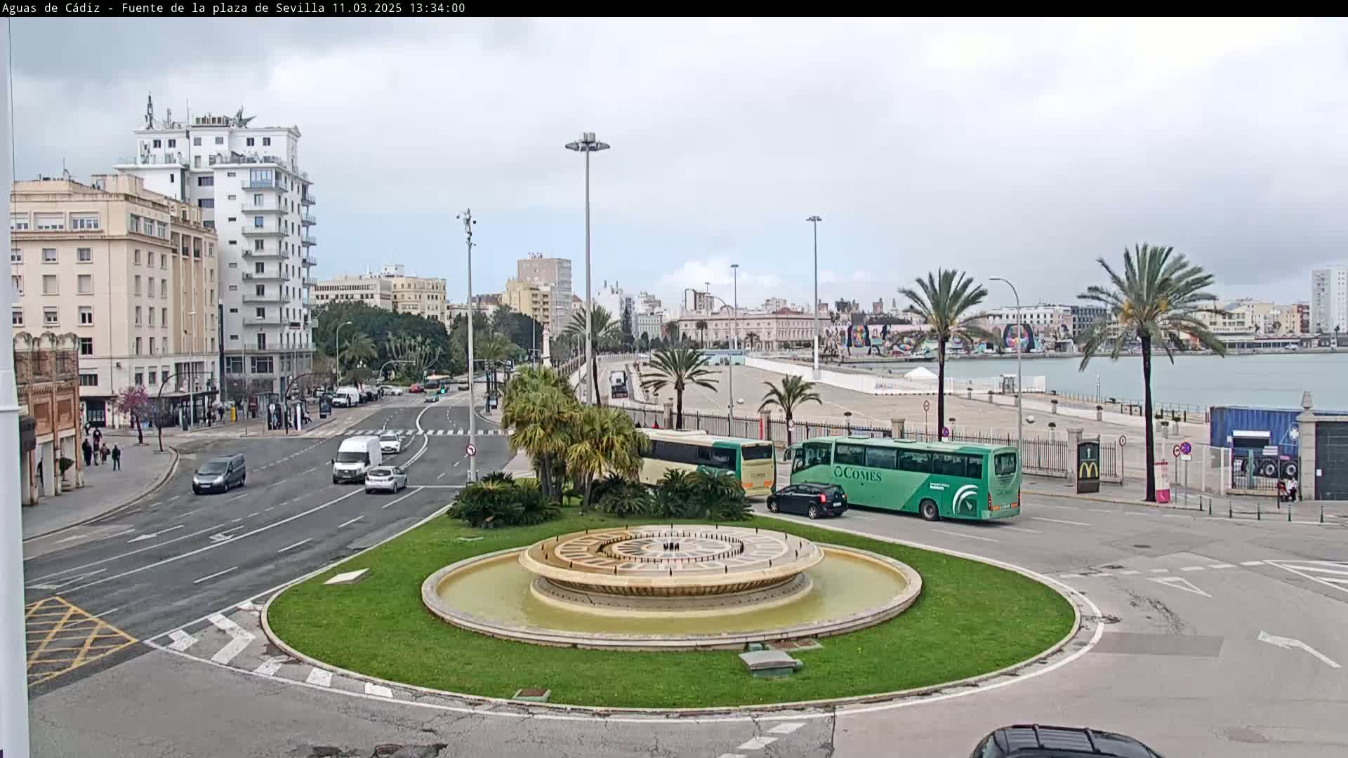 A roundabout with a circular fountain in the center, surrounded by city streets and buildings, overlooks a waterfront area under an overcast sky.