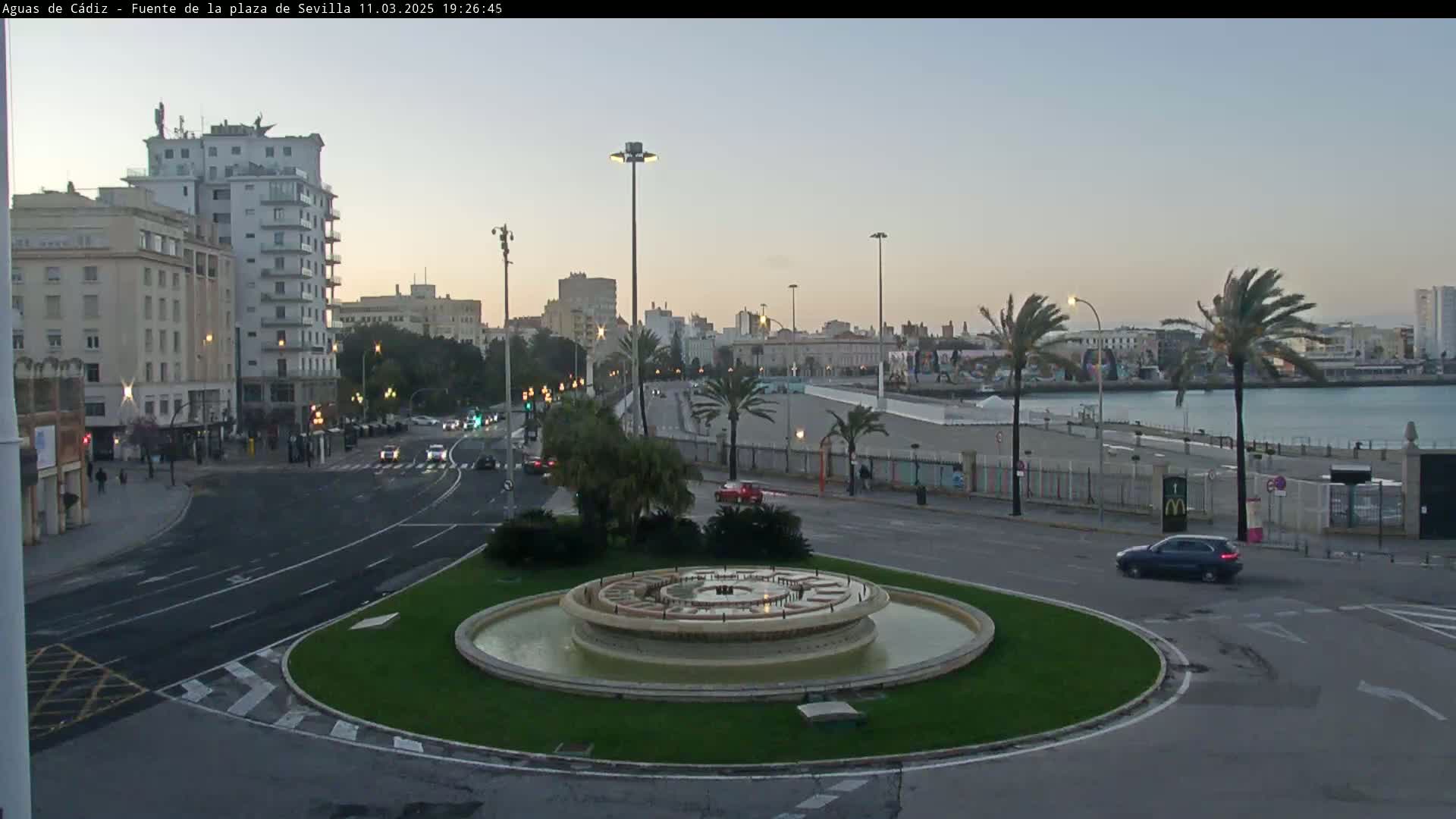A circular fountain sits in a traffic circle near a waterfront city at dusk under a clear sky.