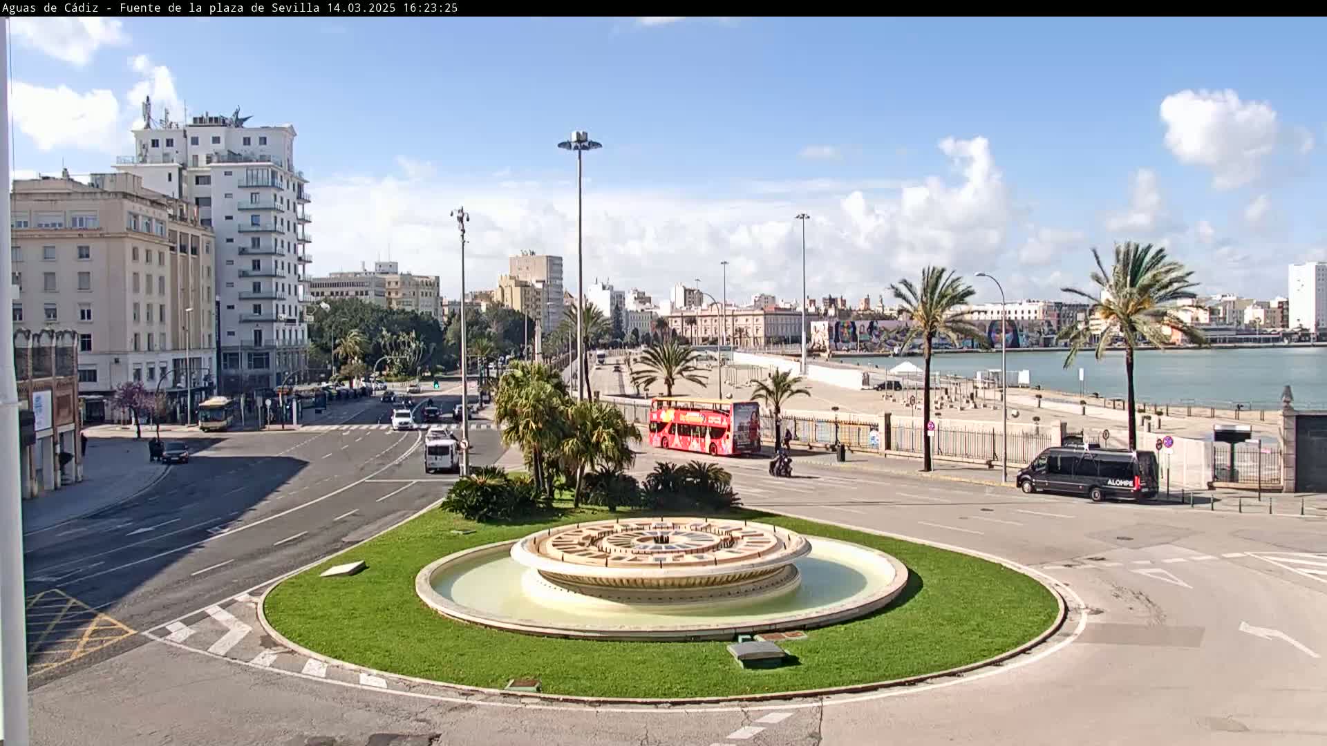 A city square with a circular fountain in the center, surrounded by roads and buildings, under a partly cloudy sky.