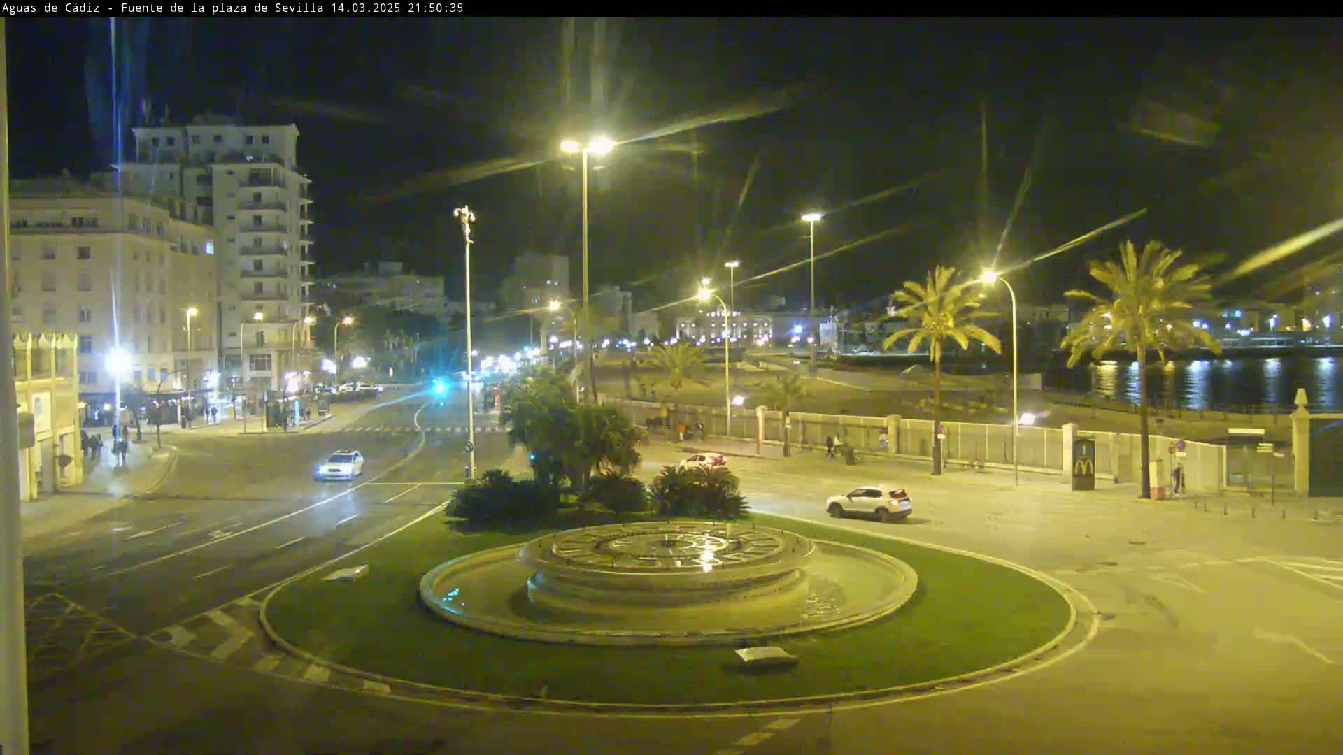 A nighttime view of a city square with a circular fountain, palm trees, and a few cars, near a body of water under clear skies.