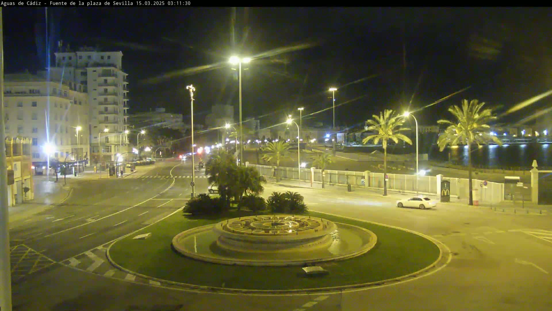 A nighttime view of a city roundabout with a fountain at its center, illuminated by streetlights, with palm trees lining a waterfront promenade in the background.