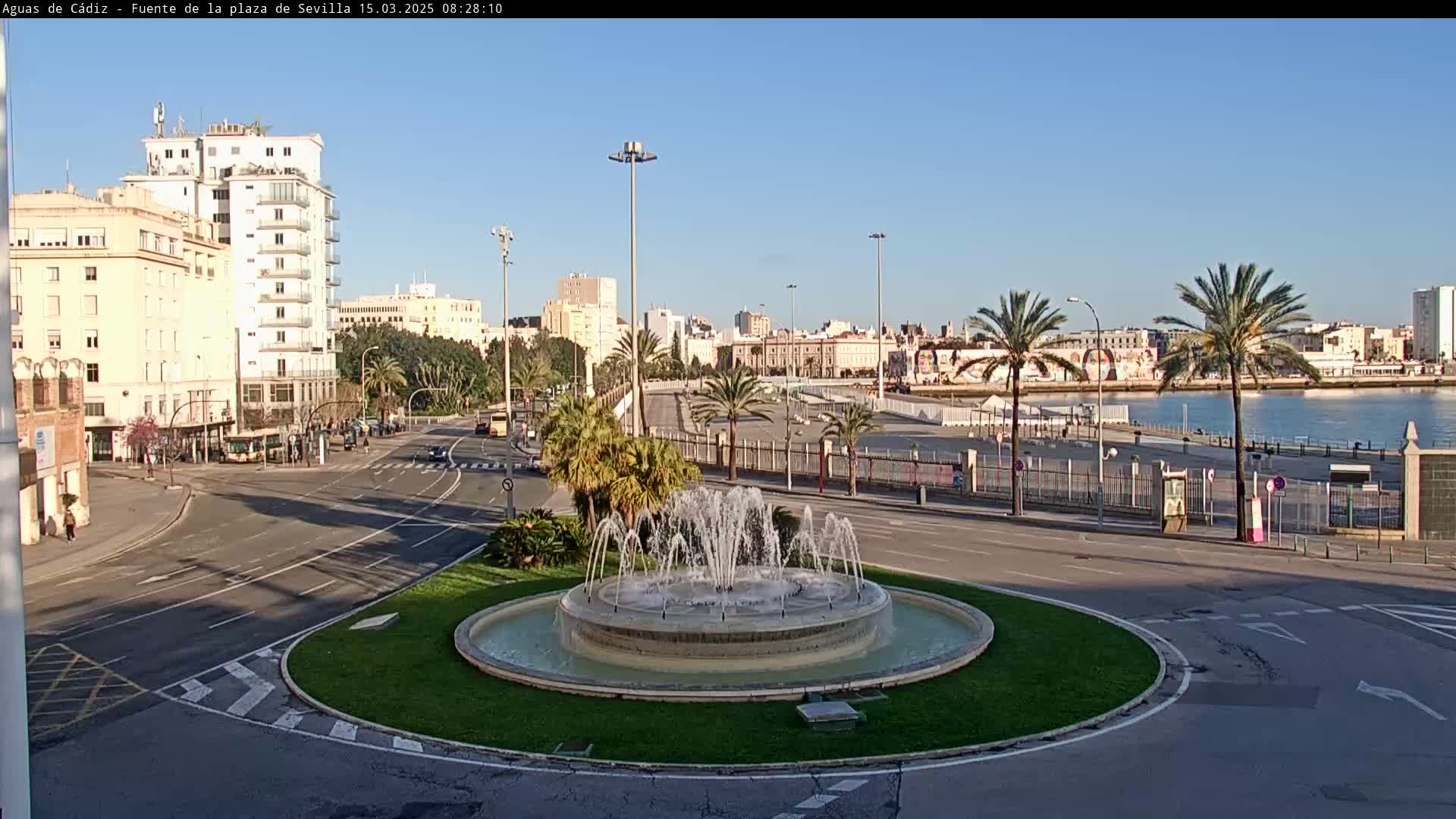 A city square with a fountain in the center, surrounded by palm trees, buildings, and a calm body of water in the background under a clear blue sky.